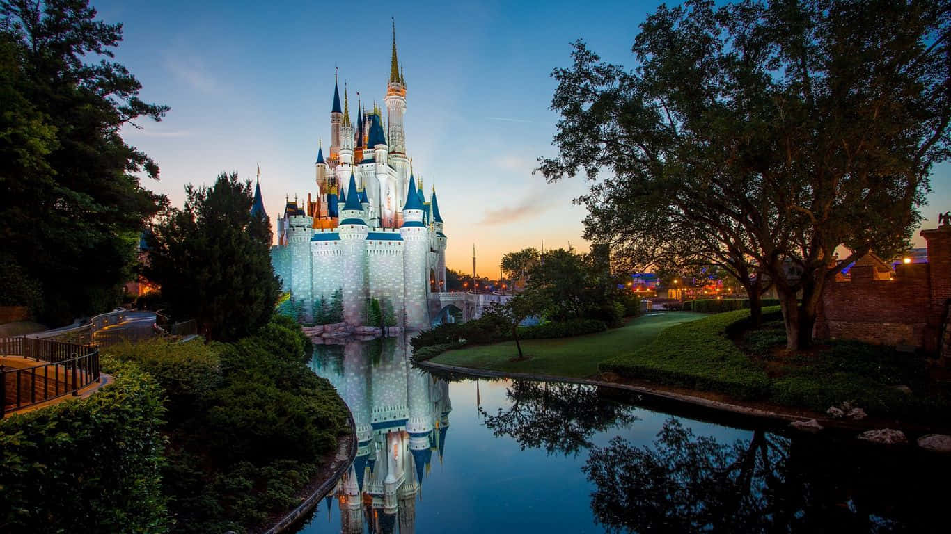 Cinderella Castle 桌布 壁紙 — 一座宏偉的迪士尼城堡在黃昏時分閃耀，其尖塔倒映在平靜的護城河中，周圍環繞著茂密的樹木。