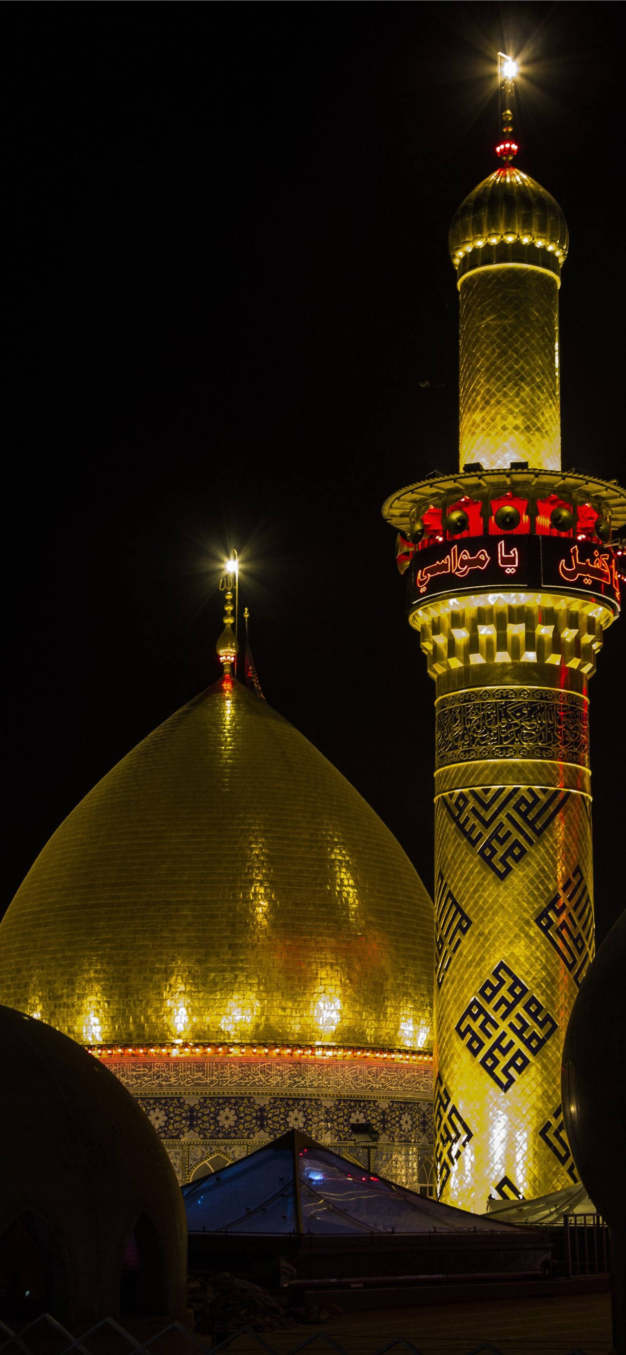 Karbala 桌布 壁紙 — 一座宏偉的金色圓頂清真寺和宣禮塔在夜空中閃耀，帶有複雜的幾何圖案和溫暖的照明。