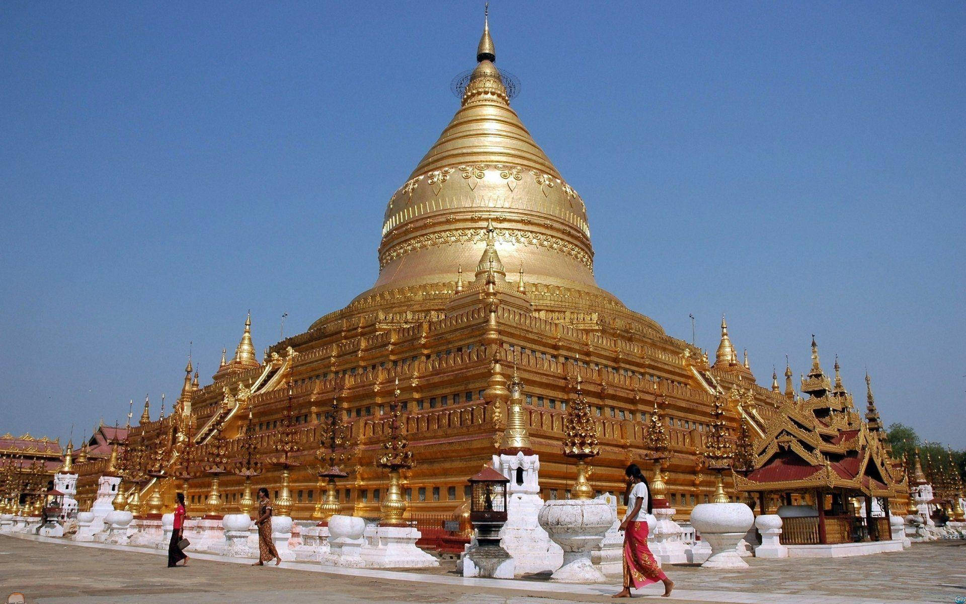 Burma 桌布 壁紙 — 一座宏偉的金色寶塔，擁有精緻的尖頂和分層屋頂，在清澈的藍天下閃閃發光，遊客沿著其白色石頭周邊行走。