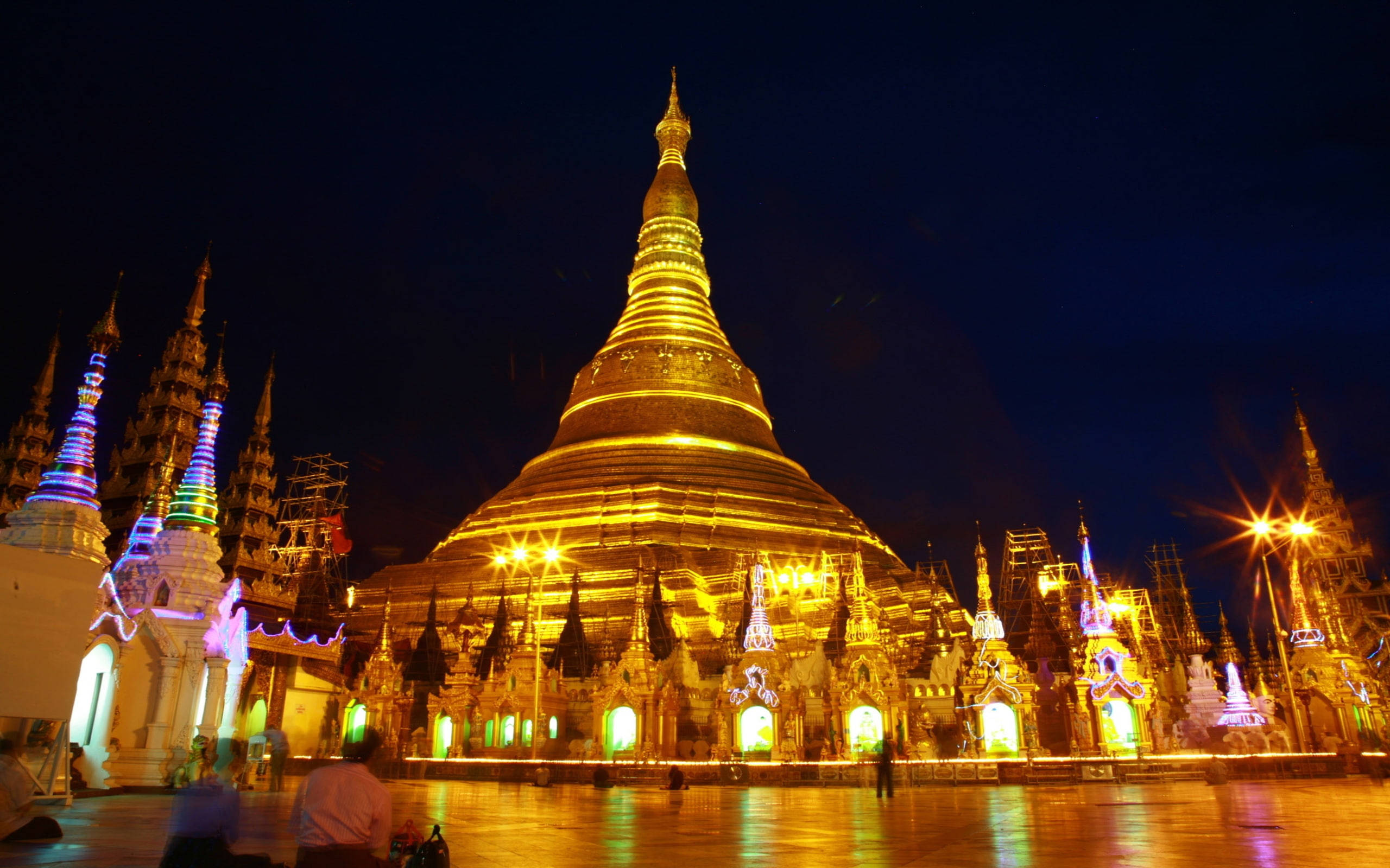 Yangon 桌布 壁紙 — 一座宏偉的金色寶塔在夜間被照亮，鮮豔的燈光在黑暗的天空下反射在潮濕的路面上。