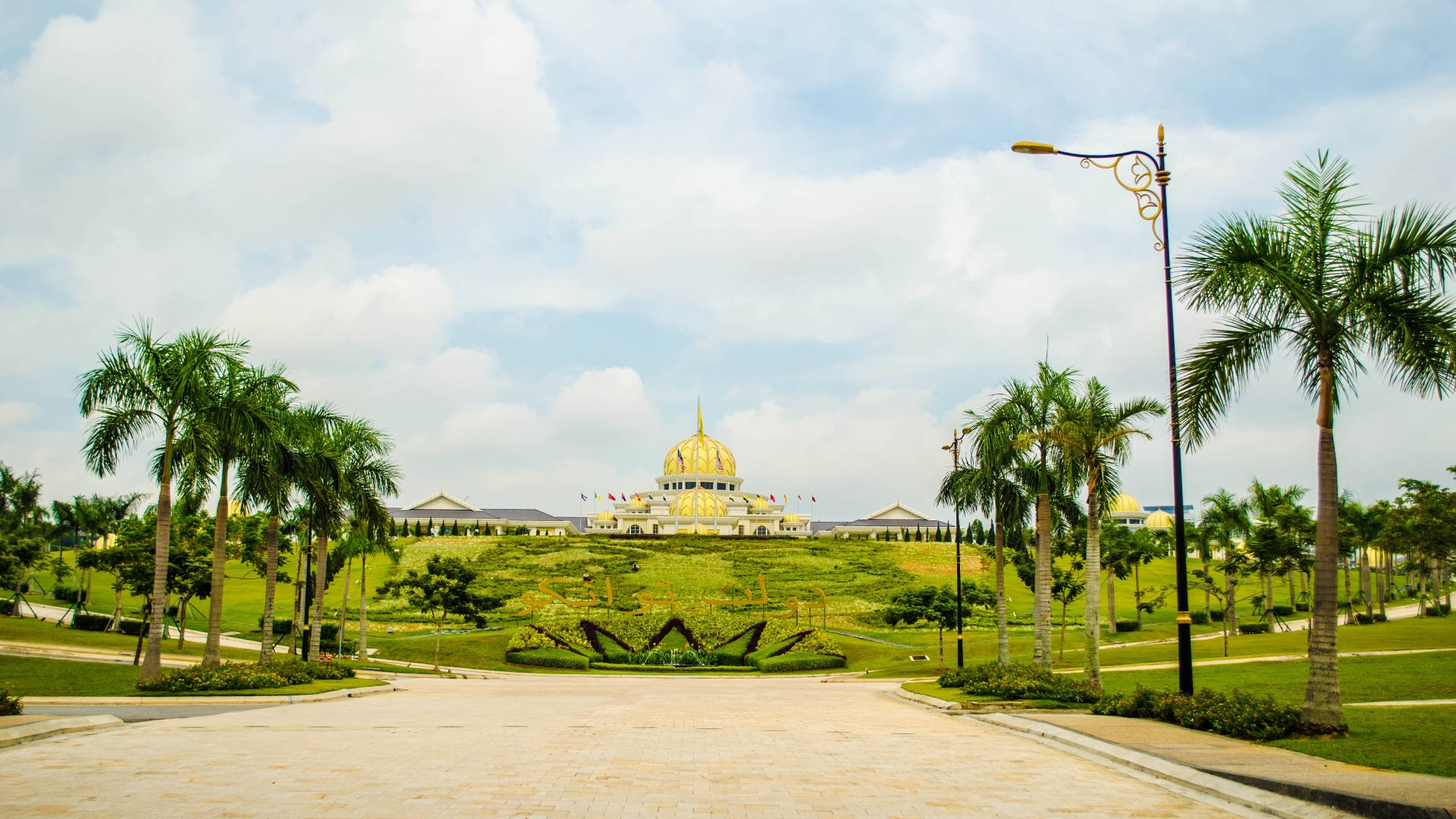 Jakarta 桌布 壁紙 — 一座宏偉的金頂清真寺坐落在鬱鬱蔥蔥的綠色山丘上，周圍環繞著棕櫚樹，天空部分多雲。