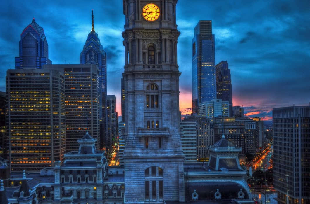 Philly 桌布 壁紙 — 一座宏偉的鐘樓在戲劇性的暮色天空下，聳立於現代摩天大樓之間，窗戶閃爍著光芒。