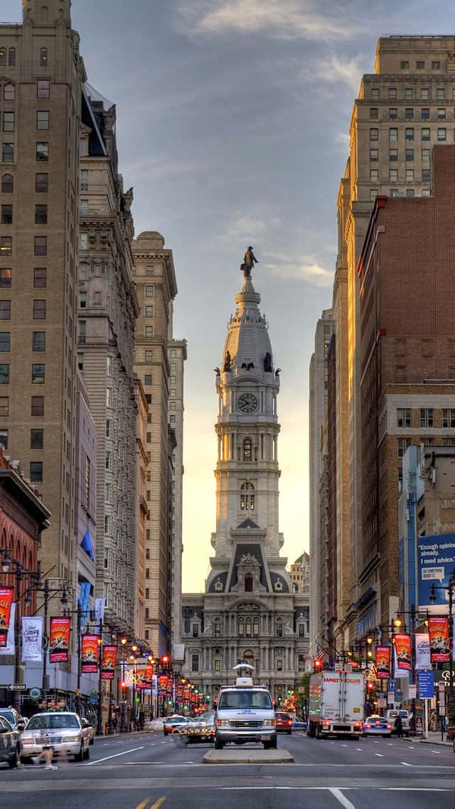 Philly 桌布 壁紙 — 一座宏偉的鐘樓在高聳的城市建築之間巍然聳立，陽光灑落在下方的城市街道上。