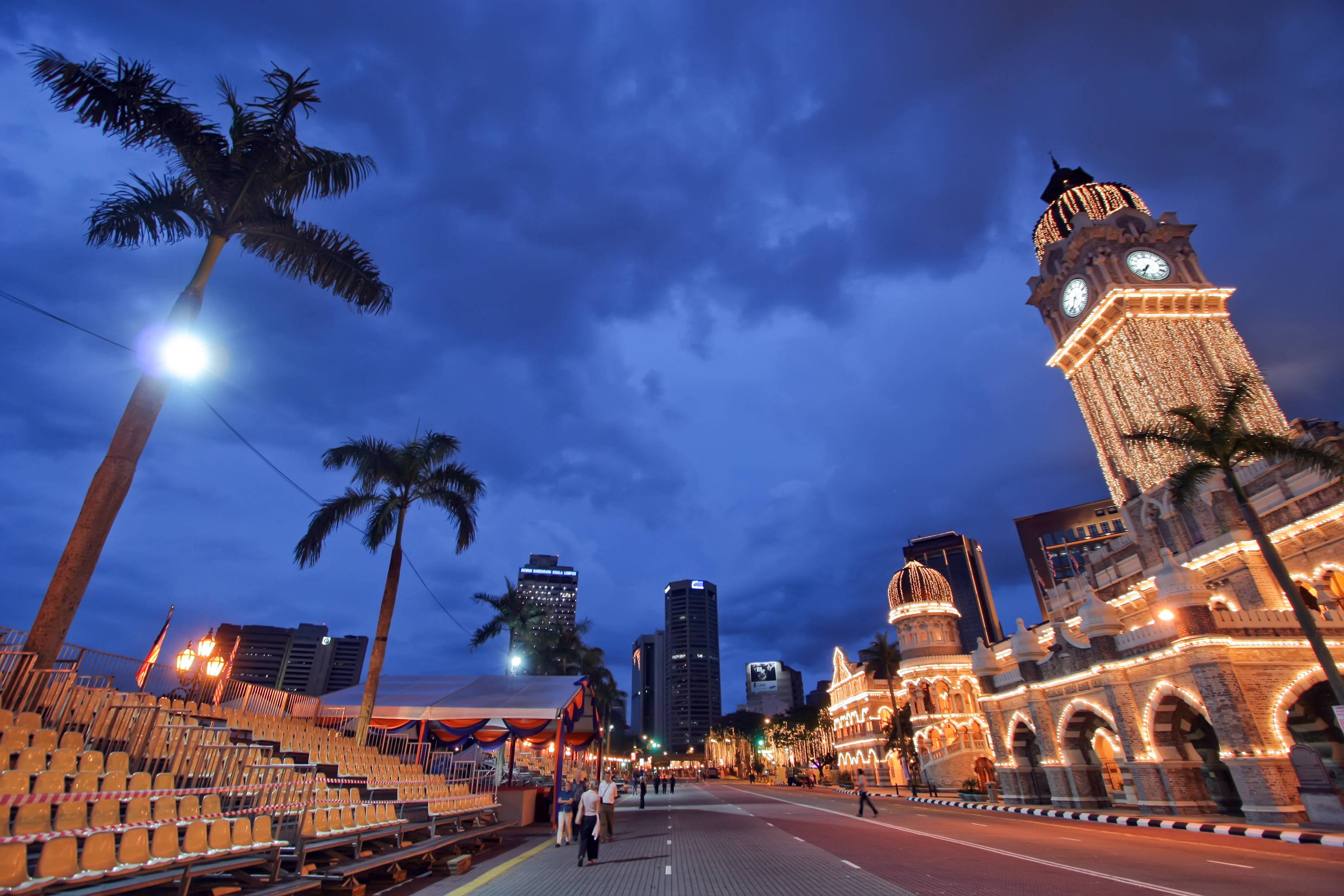Kuala 桌布 壁紙 — 一座宏偉的鐘樓點綴著金色燈光，在深藍色的暮色天空中高聳，兩旁是棕櫚樹和現代摩天大樓。