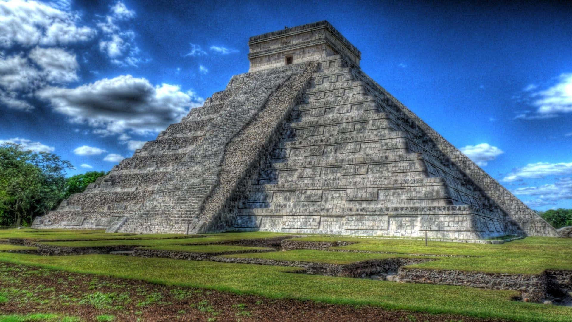 pyramid 桌布 壁紙 — 一座宏偉的階梯金字塔神廟在湛藍的天空下聳立，點綴著蓬鬆的白雲，底部是茂盛的綠草。
