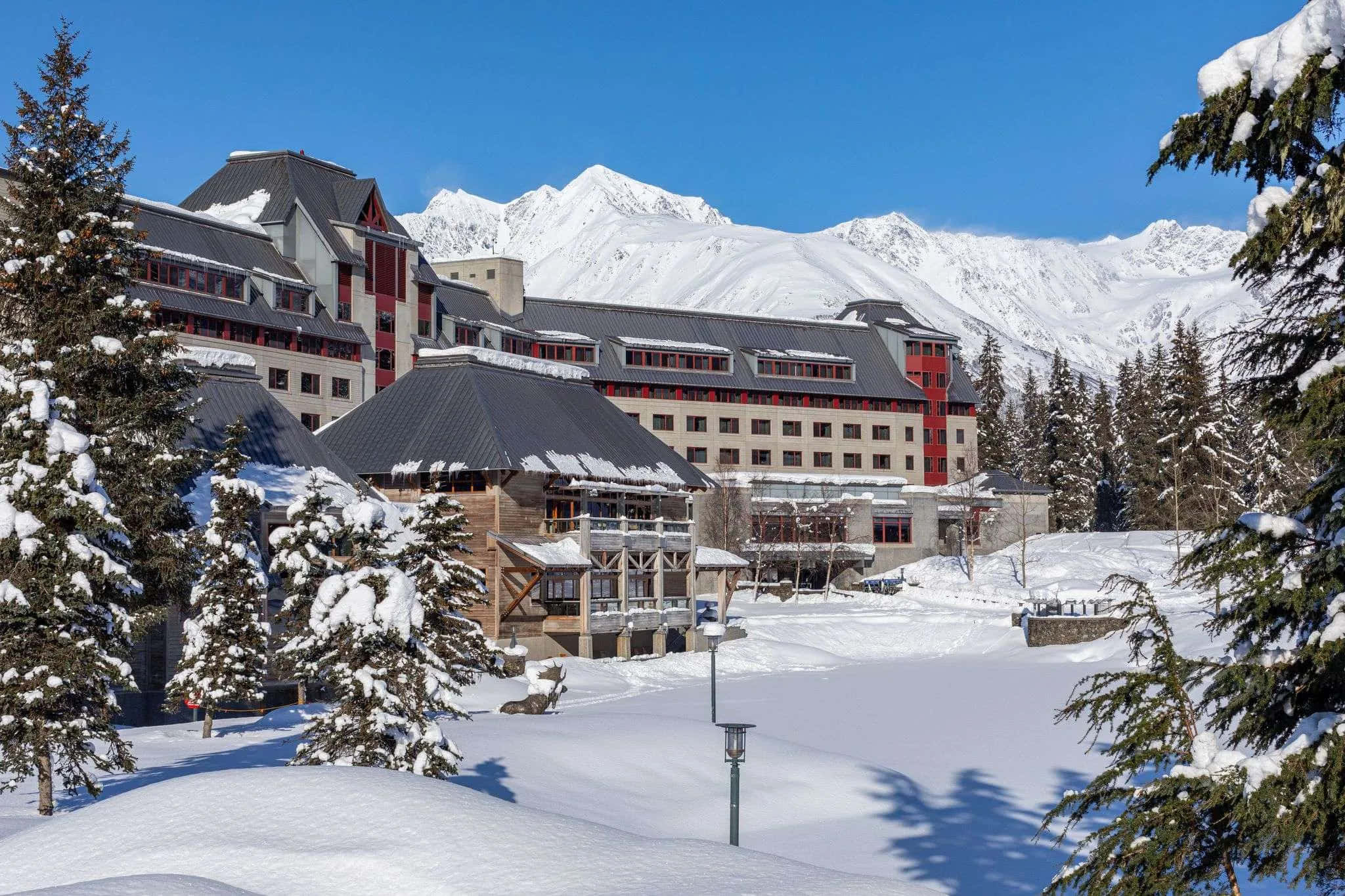 Resort 桌布 壁紙 — 一座宏偉的雪覆蓋山區度假酒店，坐落在常青樹叢中，背景是湛藍天空下高聳的山峰。