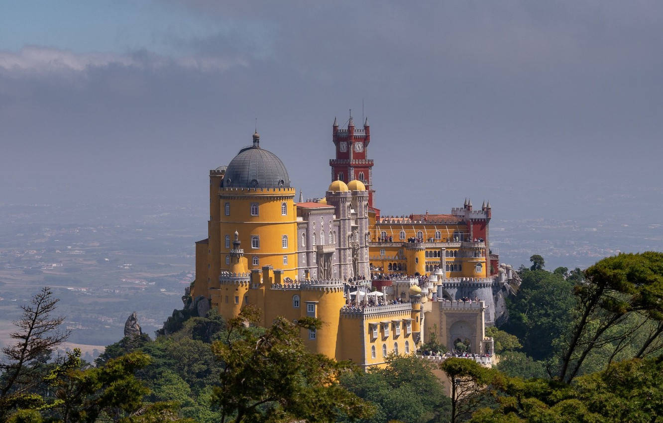 Portugal 桌布 壁紙 — 一座宏偉的黃色和紅色宮殿坐落在山頂上，周圍環繞著茂密的綠色樹木，在陰天天空下呈現朦朧的遠景。