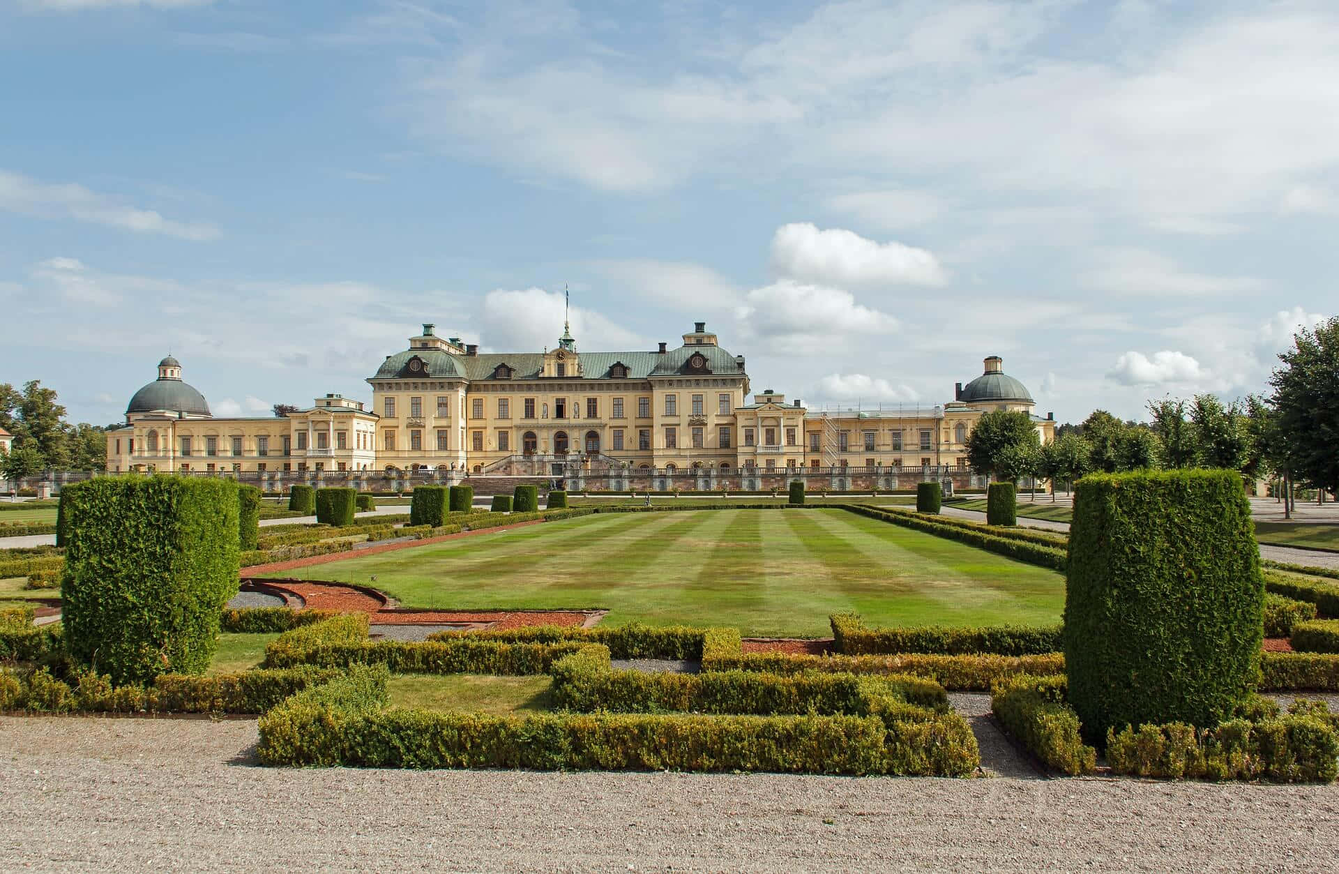 Drottningholm Palace 桌布 壁紙 — 一座宏偉的黃色宮殿，帶有綠色圓頂，矗立在修剪整齊的花園後面，上方是晴朗的藍天和白雲。