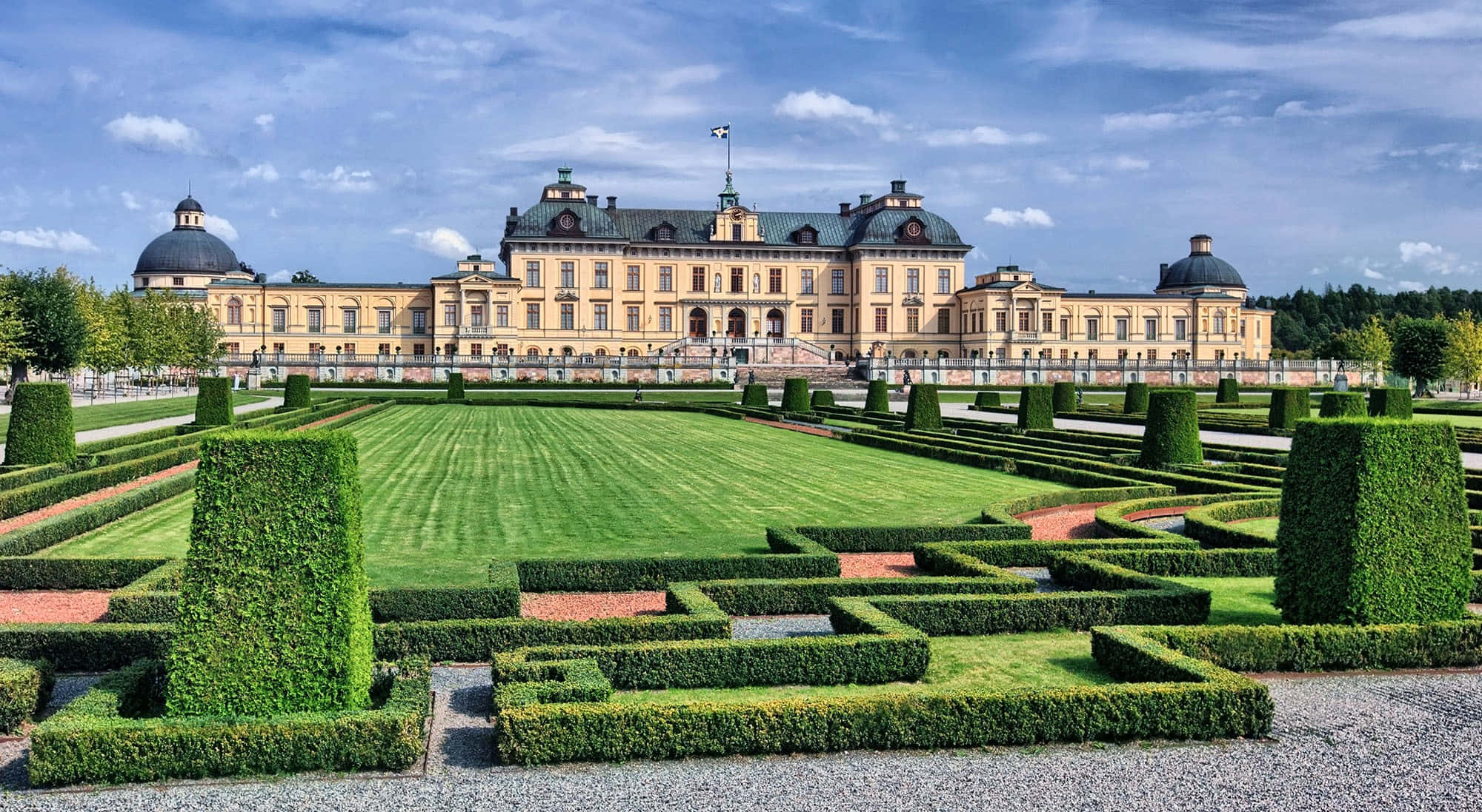 Drottningholm Palace 桌布 壁紙 — 一座宏偉的黃色宮殿，擁有圓頂塔樓，俯瞰著精心修剪的花園，園中有綠色樹籬和條紋草坪，上方是明亮的藍天。