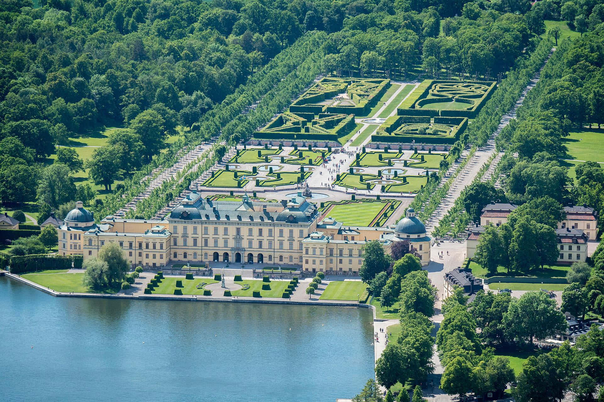 Drottningholm Palace 桌布 壁紙 — 一座宏偉的黃色宮殿的鳥瞰圖，擁有對稱的花園和一個被茂密綠樹環繞的湖泊，在晴朗的日子裡。