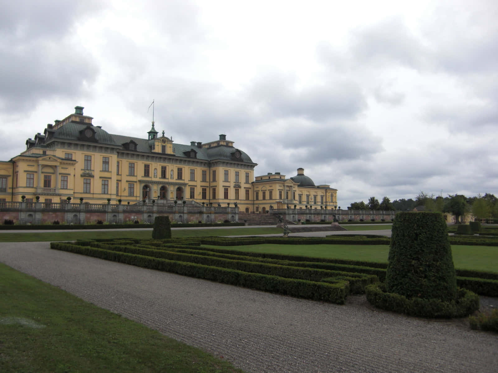 Drottningholm Palace 桌布 壁紙 — 一座宏偉的黃色巴洛克式宮殿，擁有綠色屋頂，矗立在多雲的天空下，四周環繞著精心修剪的樹籬和碎石路。