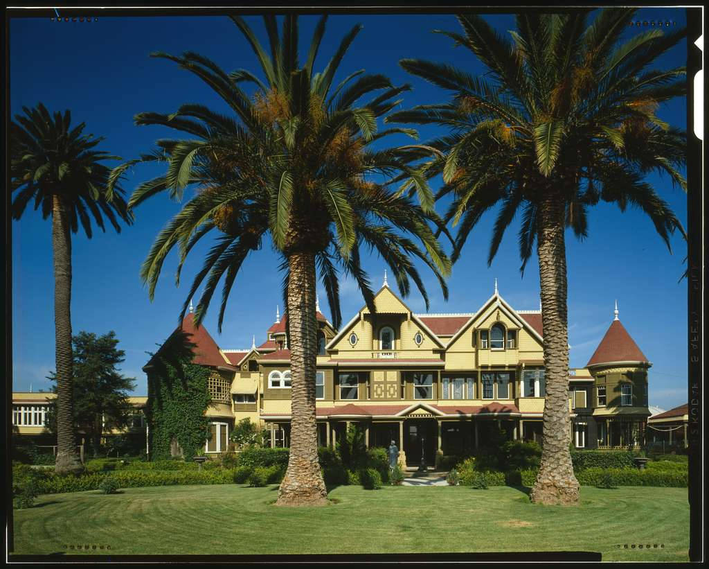 Mansion 桌布 壁紙 — 一座宏偉的黃色維多利亞式豪宅，擁有紅色屋頂和塔樓，矗立在晴朗的藍天下，周圍環繞著高大的棕櫚樹和修剪整齊的綠色草坪。