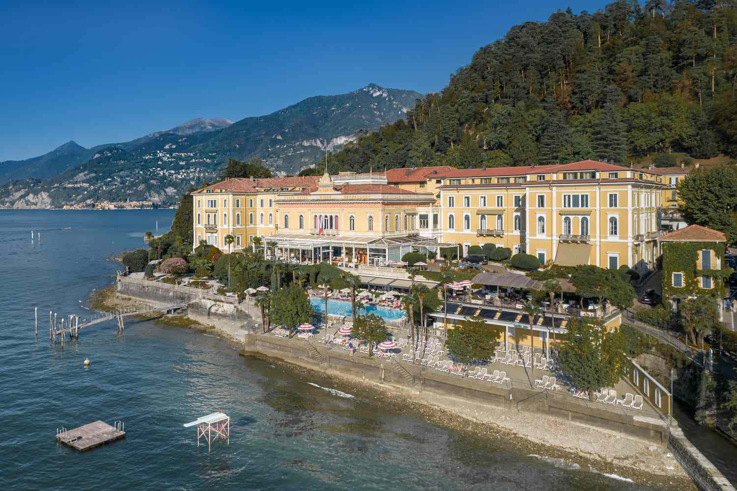 Italian 桌布 壁紙 — 一座宏偉的黃色酒店，紅色屋頂矗立在湖岸邊，四周環繞著鬱鬱蔥蔥的綠色山丘和清澈的藍色湖水，天空明亮。