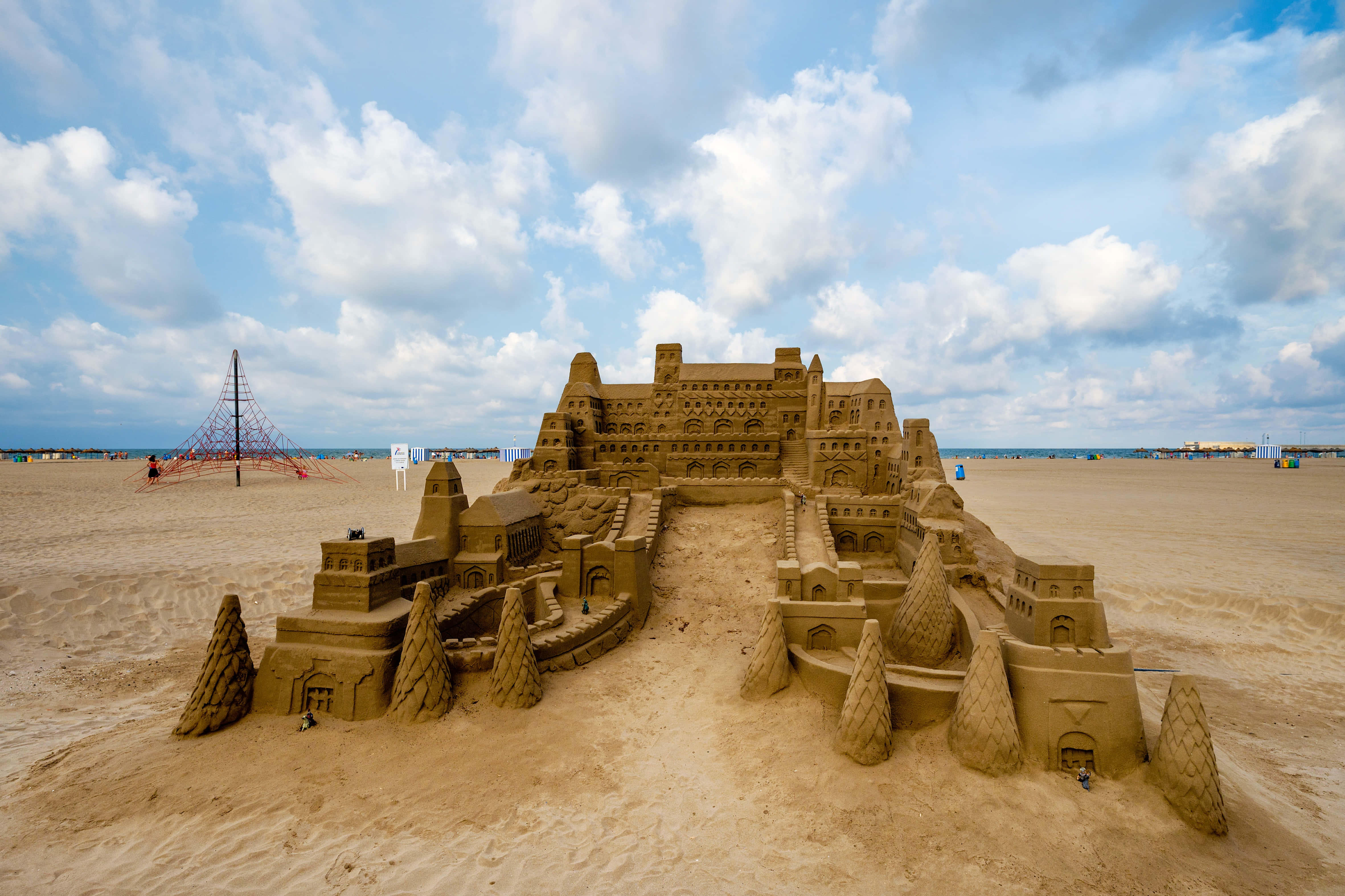 Sandcastles 桌布 壁紙 — 一座宏偉精緻的沙堡，帶有塔樓和尖頂，矗立在寬闊的海灘上，上方是明亮的藍天和蓬鬆的白雲。