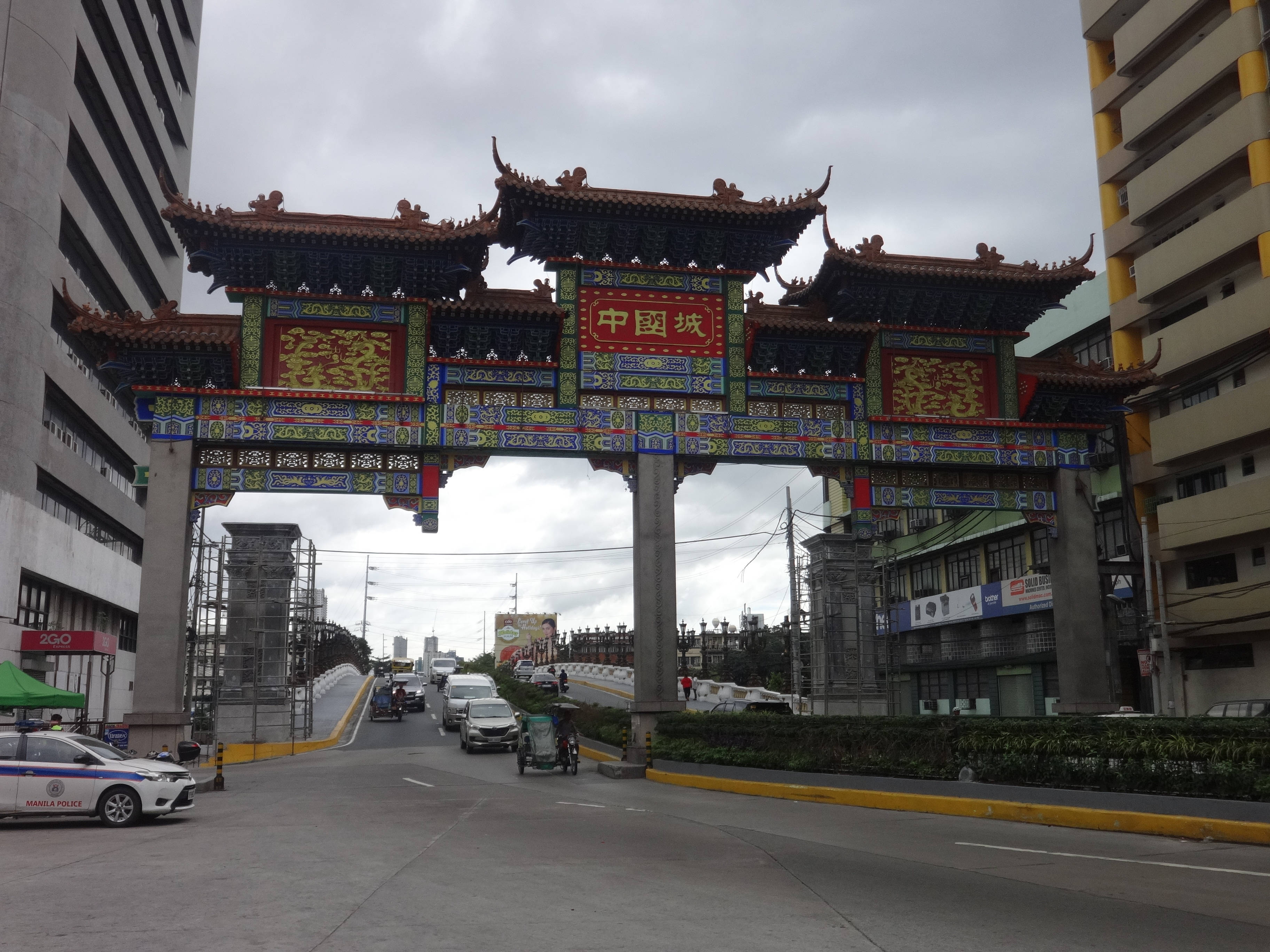 Manila 桌布 壁紙 — 一座宏偉華麗的中式牌坊，帶有紅色、金色和藍色的細節，矗立在繁忙的城市街道上，街道上有汽車和建築物，天空多雲。