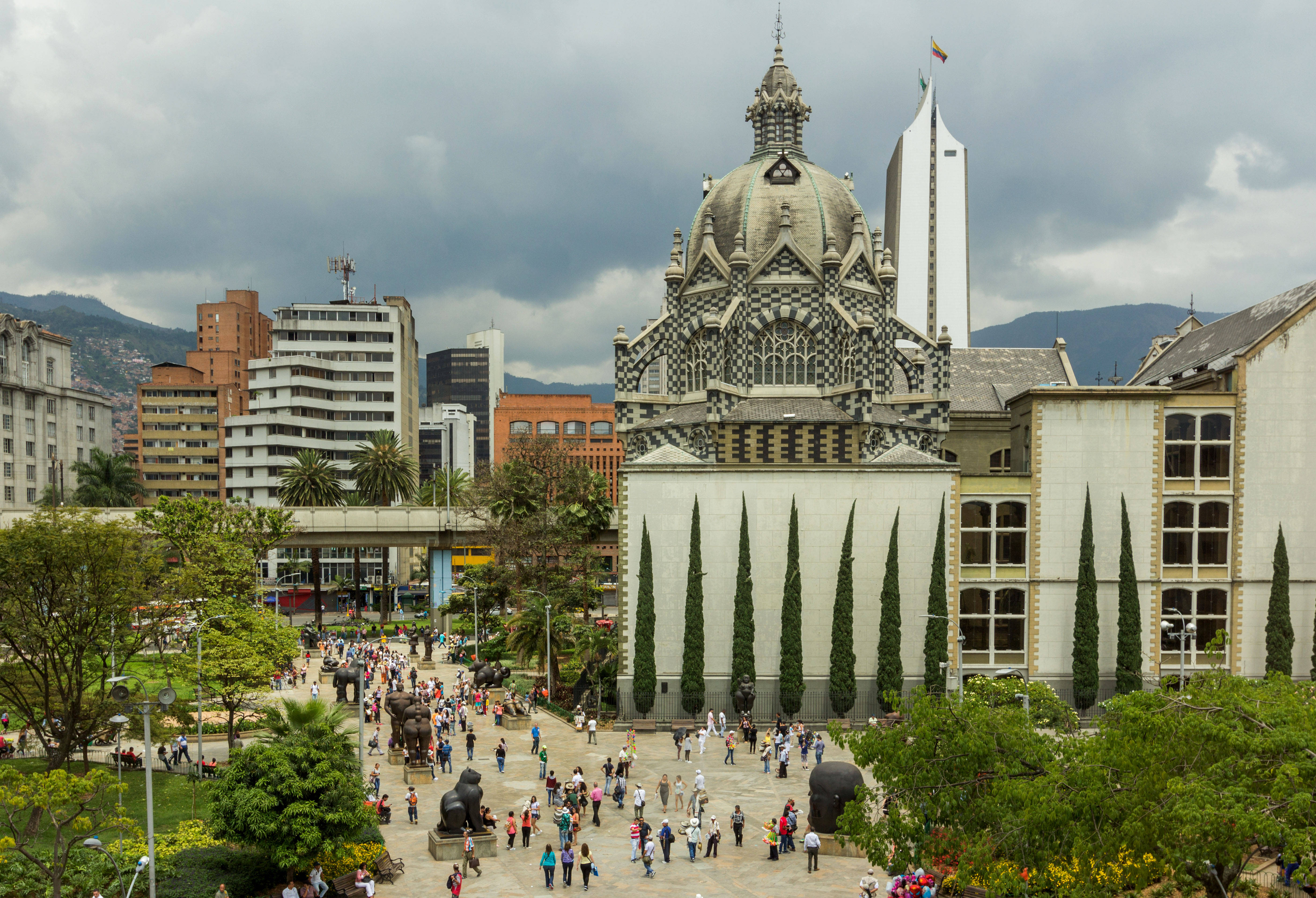 Medellin 桌布 壁紙 — 一座宏偉華麗的大教堂，帶有格子外牆，矗立在繁忙的城市廣場中，廣場上擠滿了人，兩旁是高聳的柏樹，天空多雲。
