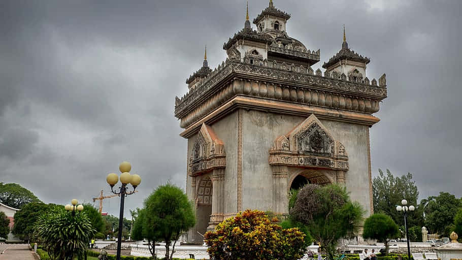 Laos 桌布 壁紙 — 一座宏偉華麗的寺廟大門矗立在沉重的灰色天空下，周圍環繞著茂密的綠樹和盛開的花朵。
