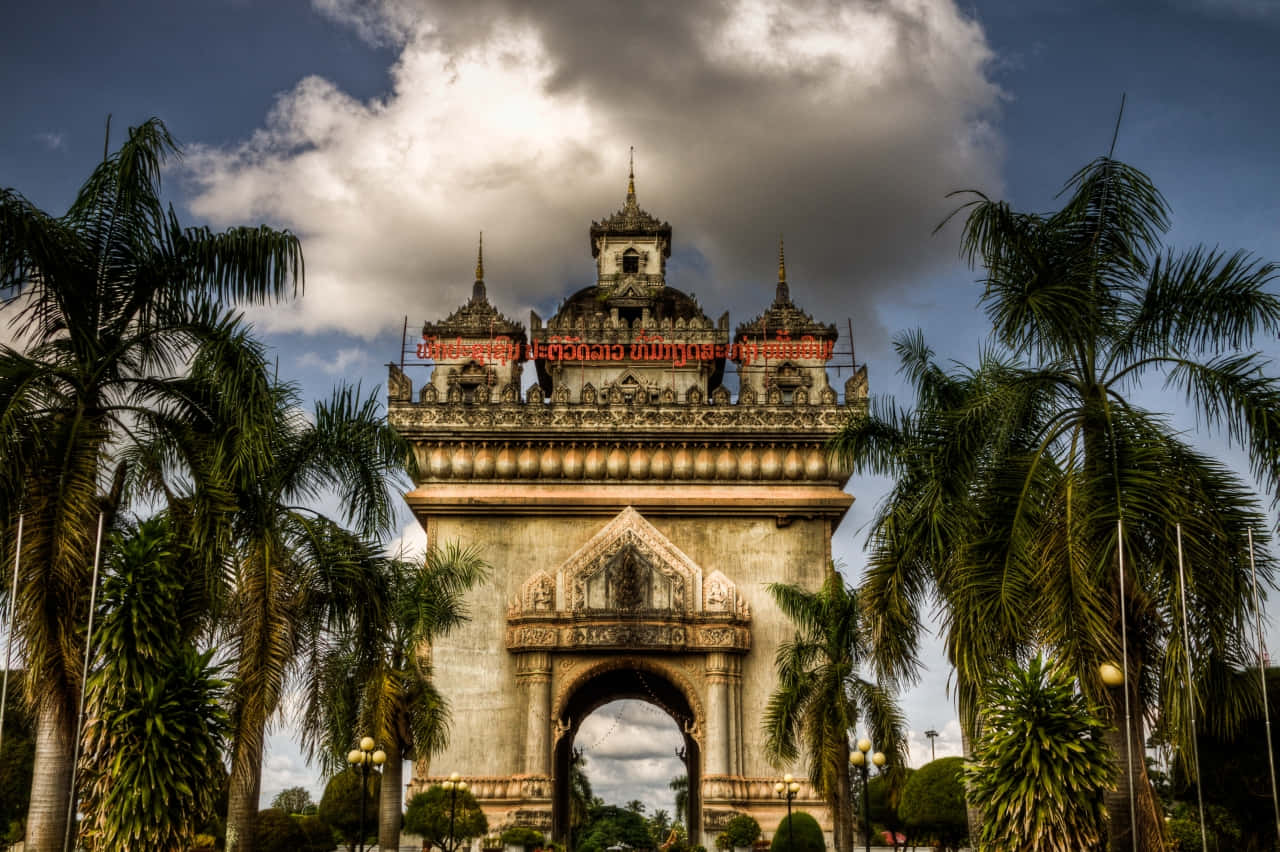 Laos 桌布 壁紙 — 一座宏偉華麗的拱門，帶有精緻雕刻和紅色文字，在戲劇性的多雲天空下高高聳立，周圍環繞著茂密的棕櫚樹。