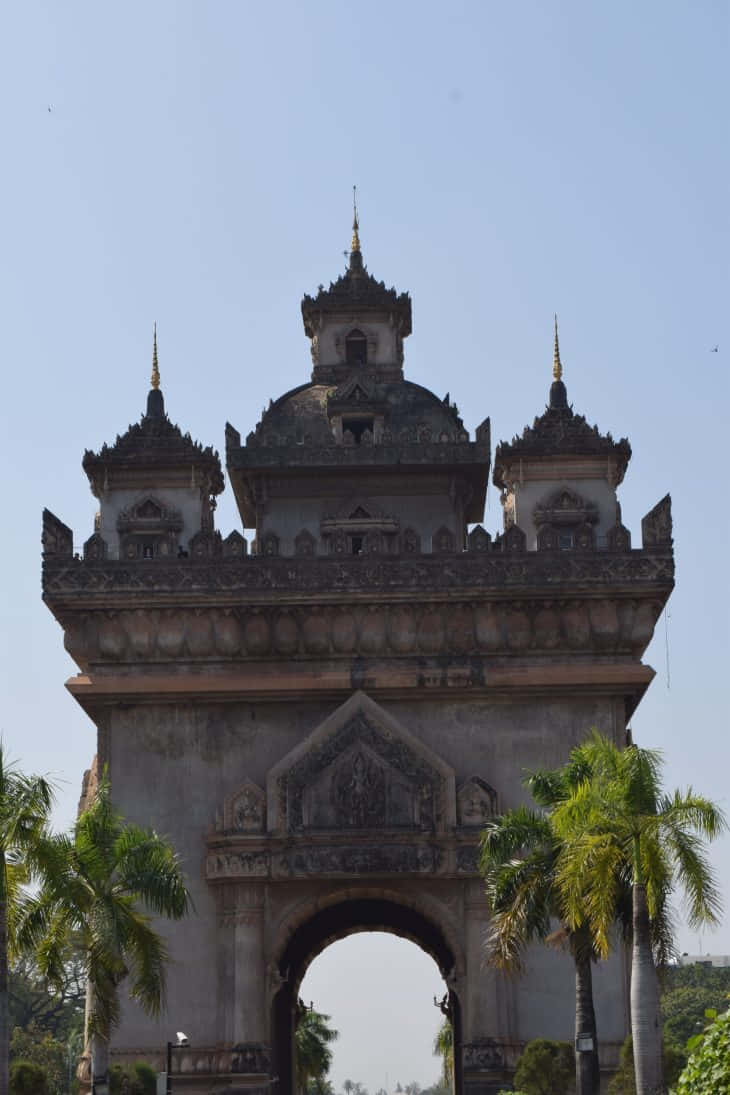 Laos 桌布 壁紙 — 一座宏偉華麗的石拱門，帶有分層尖頂和複雜雕刻，在晴朗的藍天下高高聳立，兩旁種有棕櫚樹。