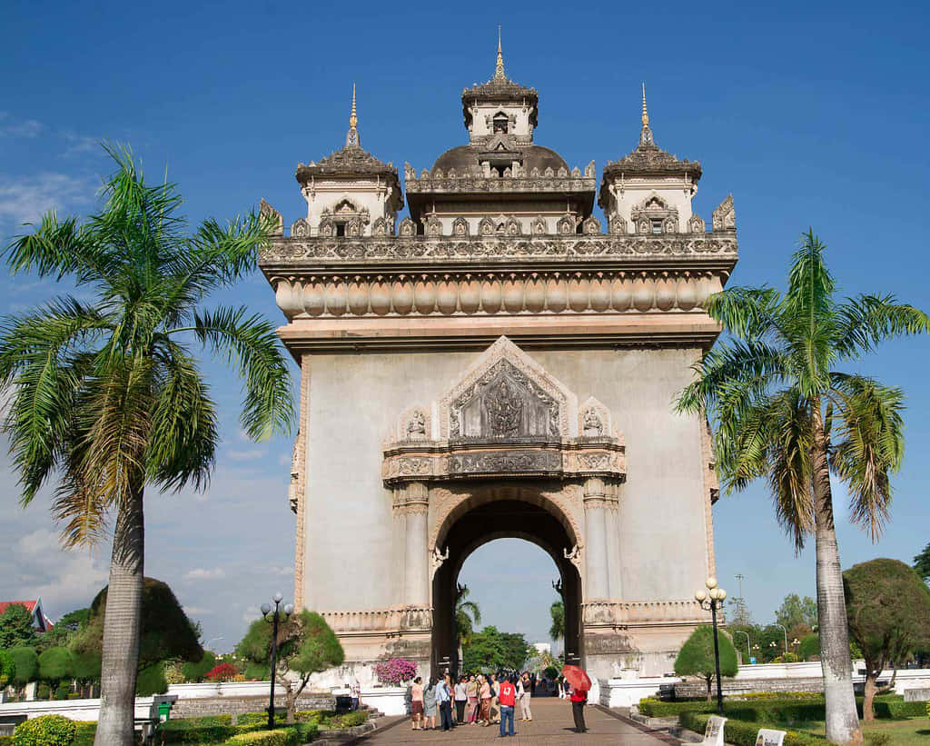 Laos 桌布 壁紙 — 一座宏偉華麗的石拱門，帶有尖塔，矗立在晴朗的藍天下，兩旁是高高的棕櫚樹和遊客。