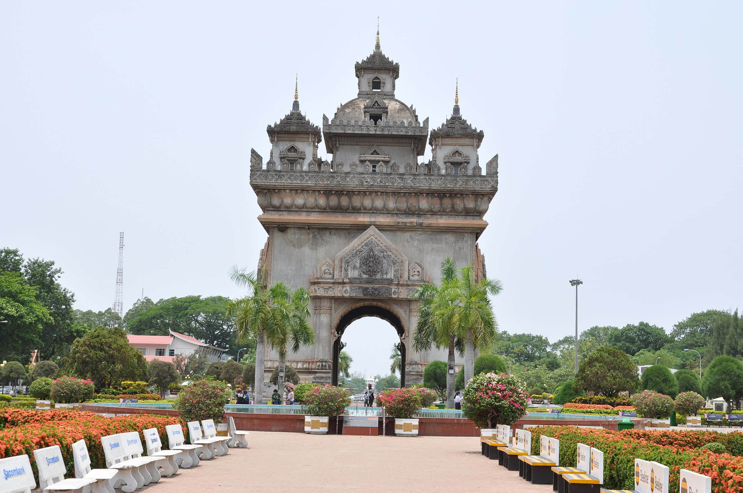 Laos 桌布 壁紙 — 一座宏偉華麗的石拱門，頂部有尖塔，矗立在鋪砌小徑的盡頭，小徑兩旁是色彩繽紛的花壇和棕櫚樹，天空多雲。