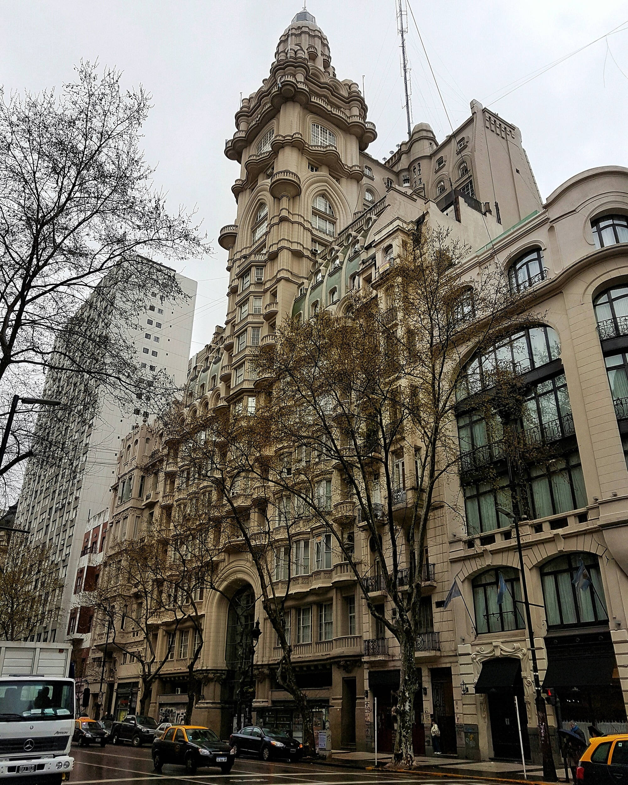 Aires 桌布 壁紙 — 一座宏偉華麗的米色建築，帶有高聳的塔樓，矗立在光禿禿的樹木和現代摩天大樓之間，天空多雲。