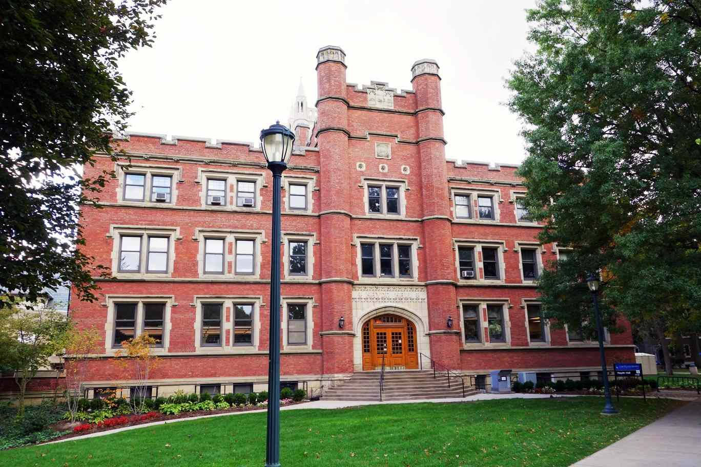 建築 桌布 壁紙 — 一座宏大的紅磚大學建築，帶有塔樓和拱形木門，矗立在綠色的草坪和秋樹之間，天空多雲。