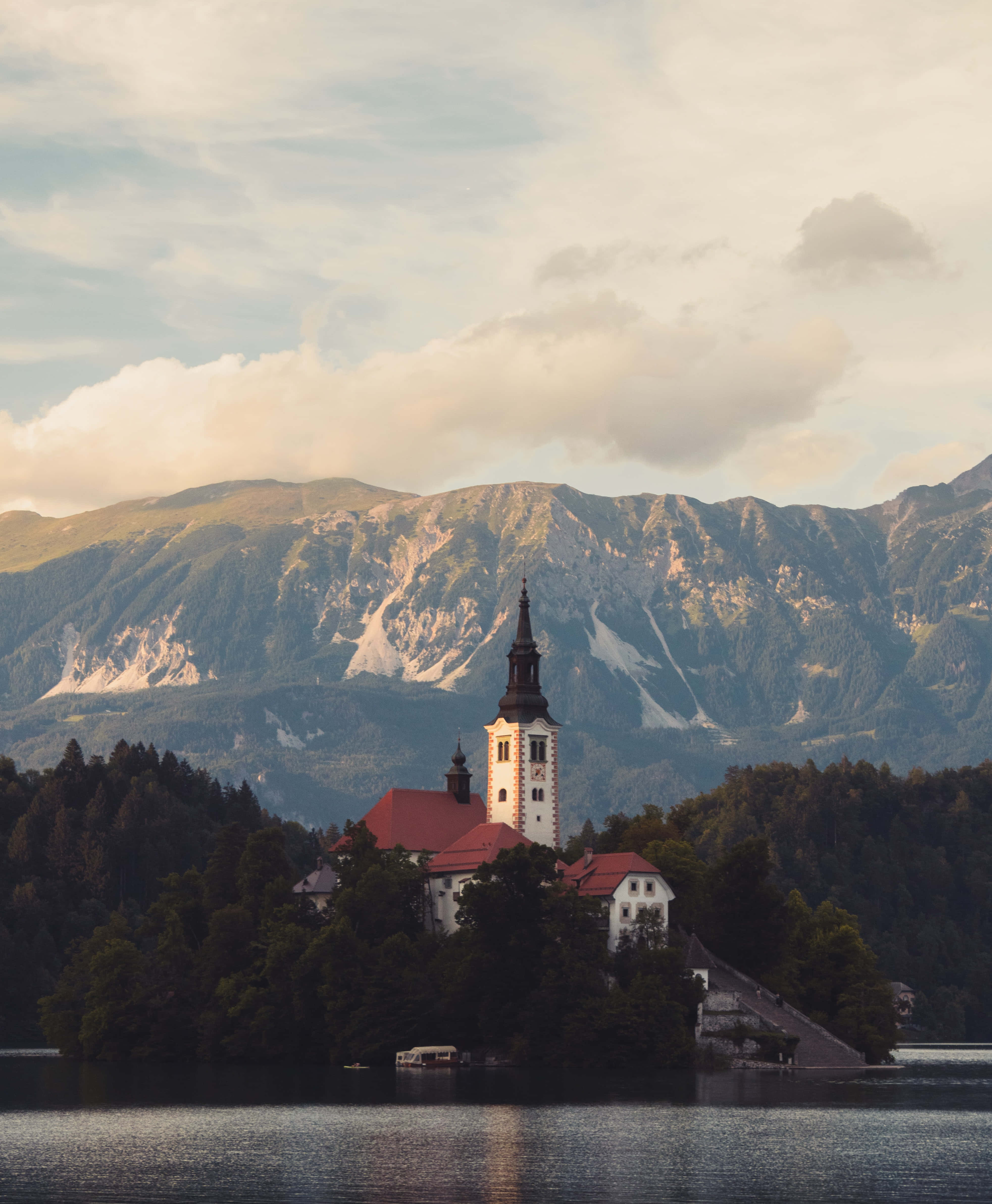 Slovenia 桌布 壁紙 — 一座寧靜的教堂，擁有高聳的尖塔，矗立在平靜湖泊中的一個樹木繁茂的島嶼上，背後是雄偉的山脈，天空柔和多雲。