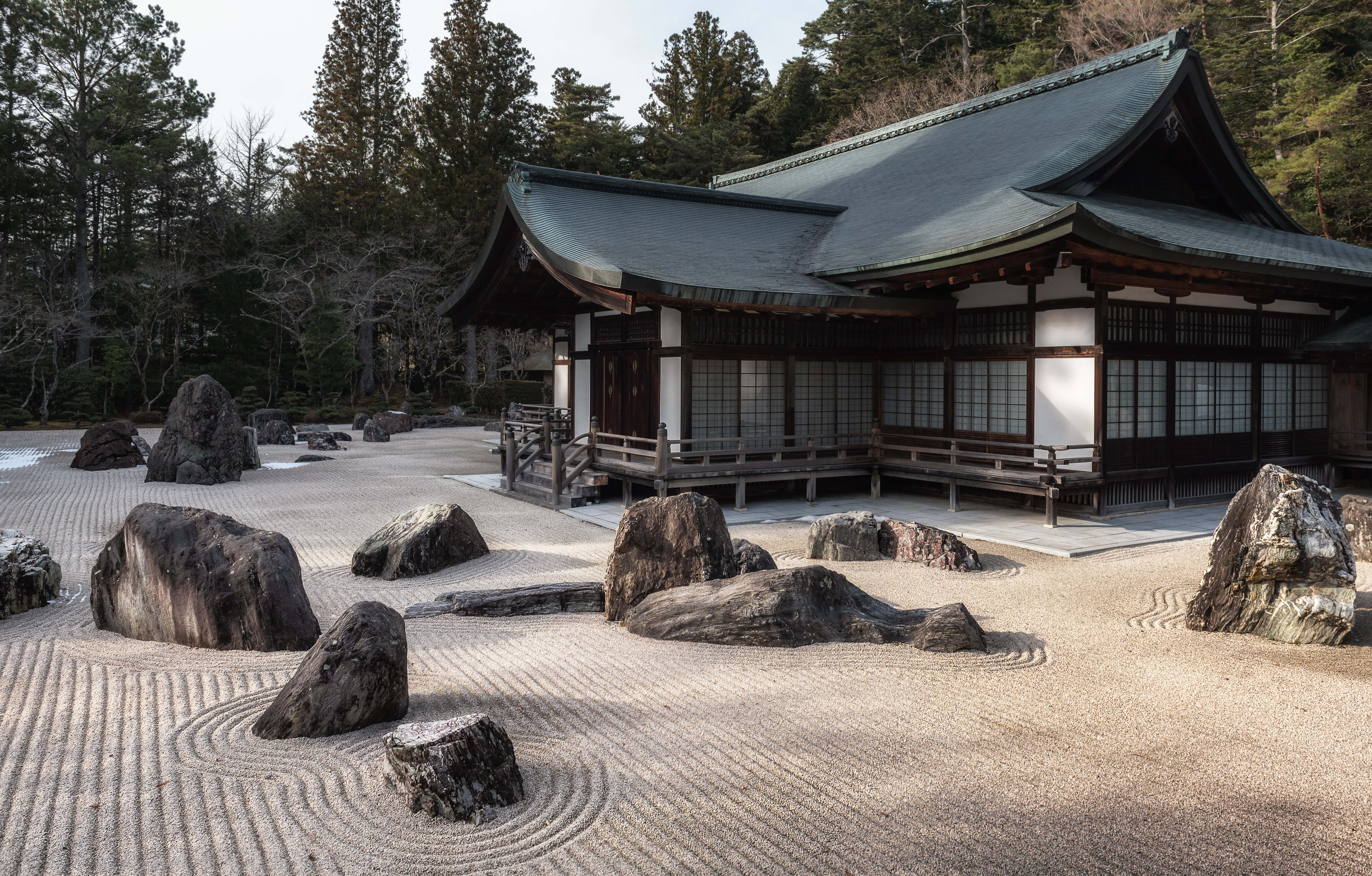 Japan Landscape 桌布 壁紙 — 一座寧靜的日本寺廟，擁有精心耙平的礫石花園和巨大的岩石，周圍環繞著高大的松樹。