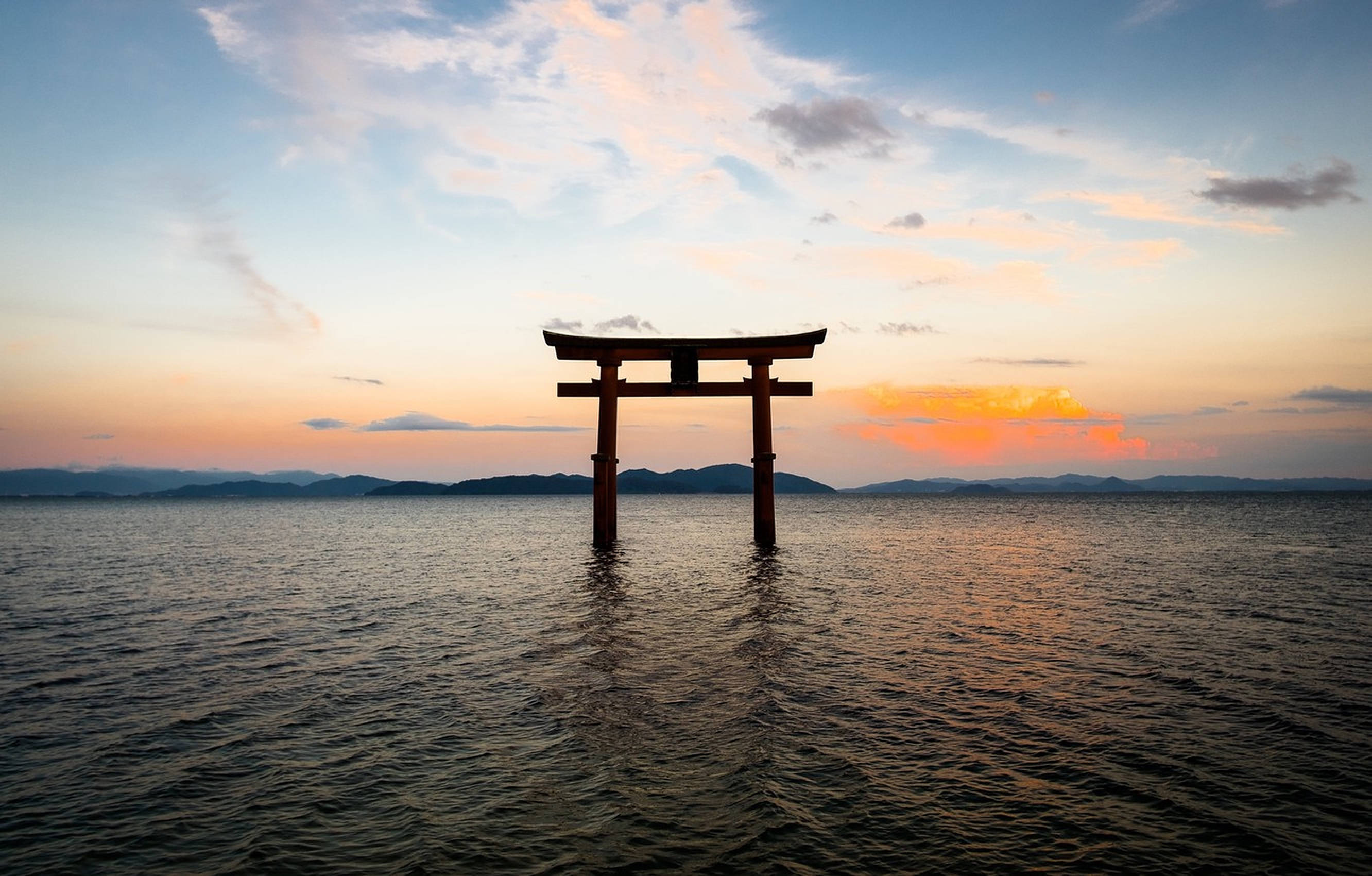 Torii 桌布 壁紙 — 一座寧靜的木製鳥居矗立在平靜的水面上，夕陽西下，橙色的雲彩和遠山映襯在柔和的藍天下。