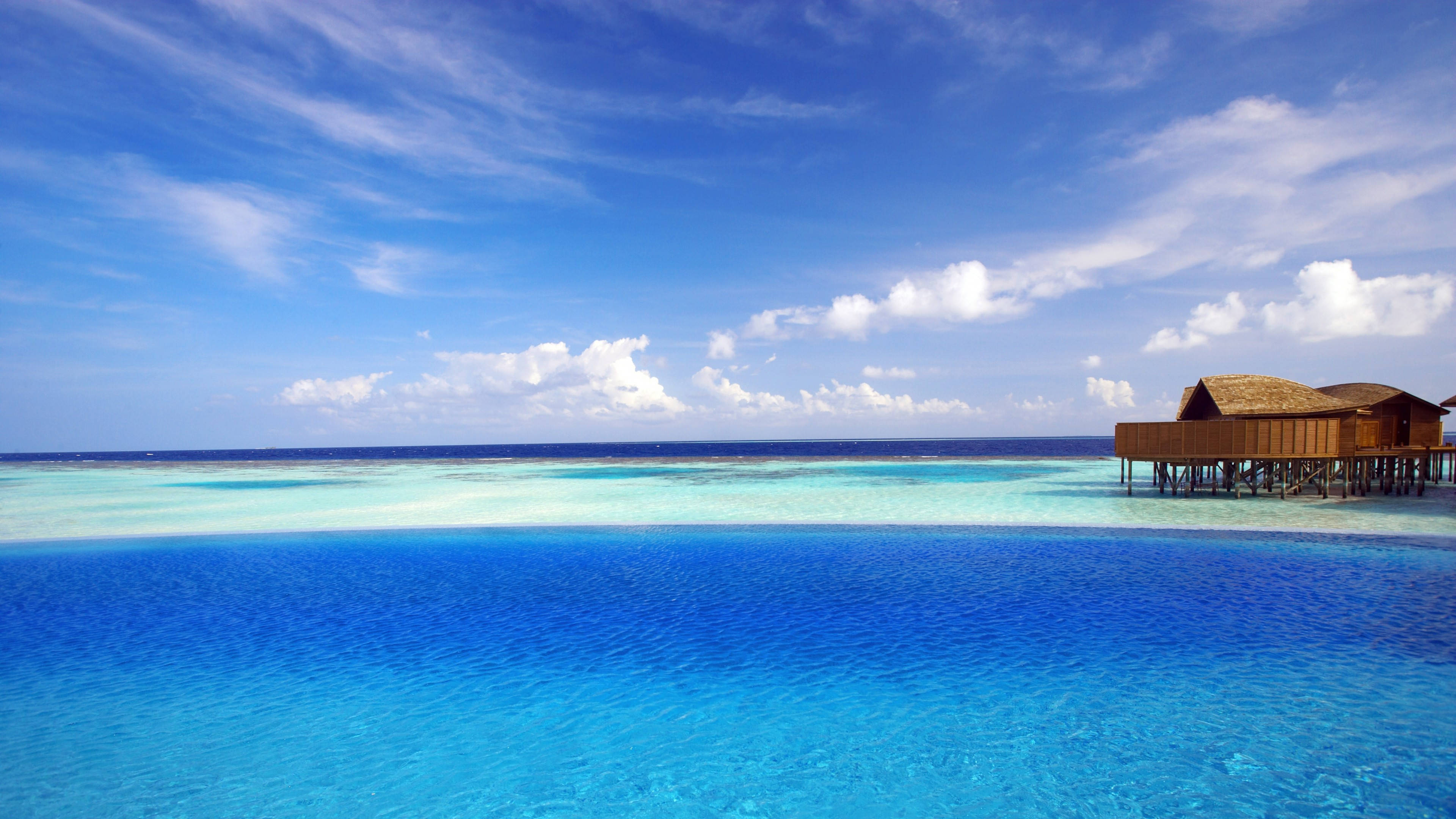 Windows 11 4K 桌布 壁紙 — 一座寧靜的水上平房，矗立在清澈碧綠的海水上的高蹺上，廣闊的藍天白雲。