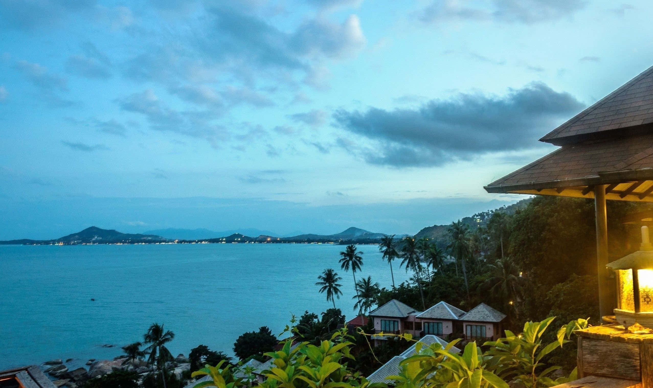 Thailand Beach 桌布 壁紙 — 一座寧靜的熱帶別墅在黃昏時分俯瞰平靜的碧綠大海，棕櫚樹和遠山在柔和的藍天下。