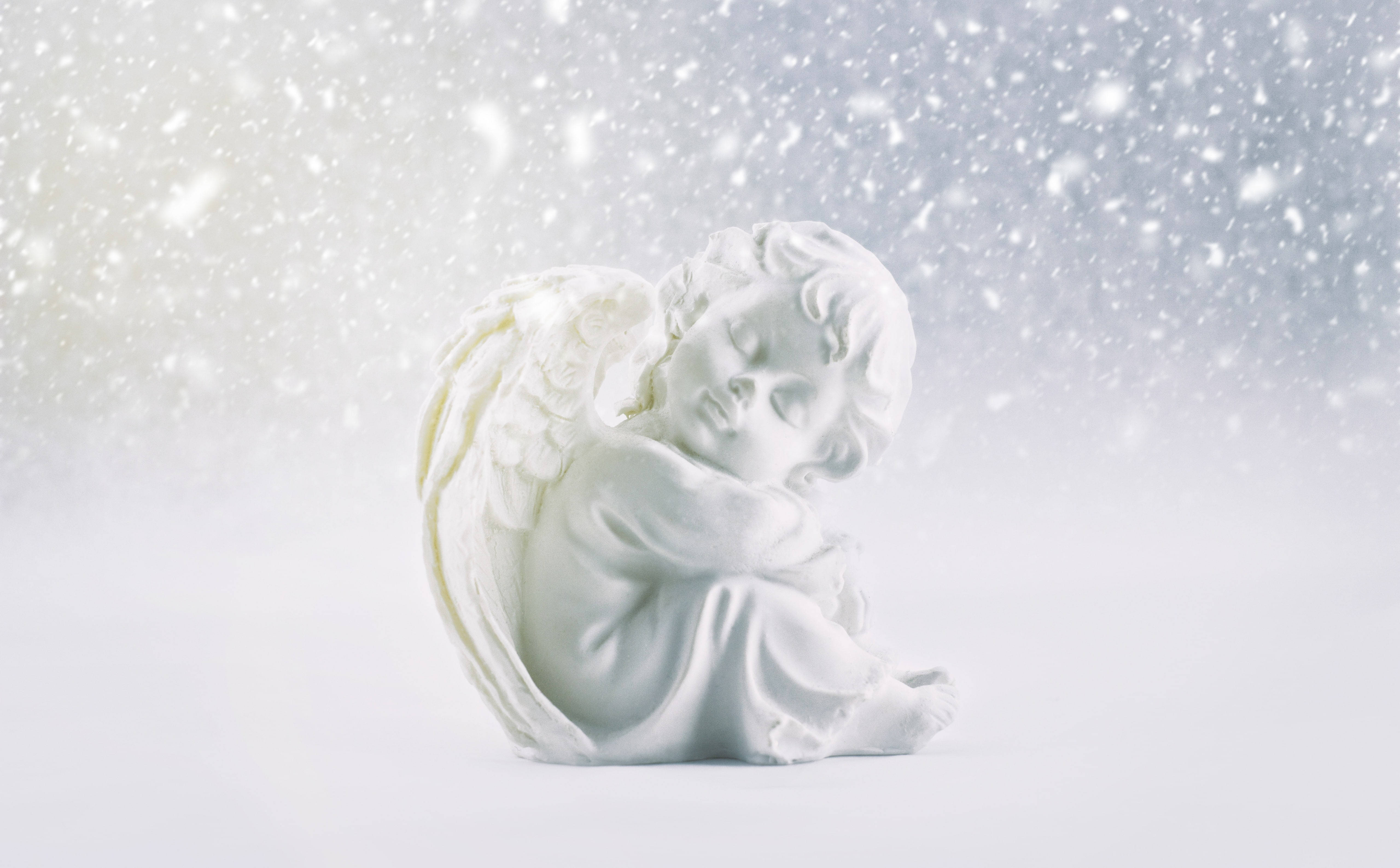 Cute Winter 桌布 壁紙 — 一座寧靜的白色大理石天使雕像蜷縮在安詳的睡眠中，溫柔的雪花在柔和發光的冬日天空中飄舞。