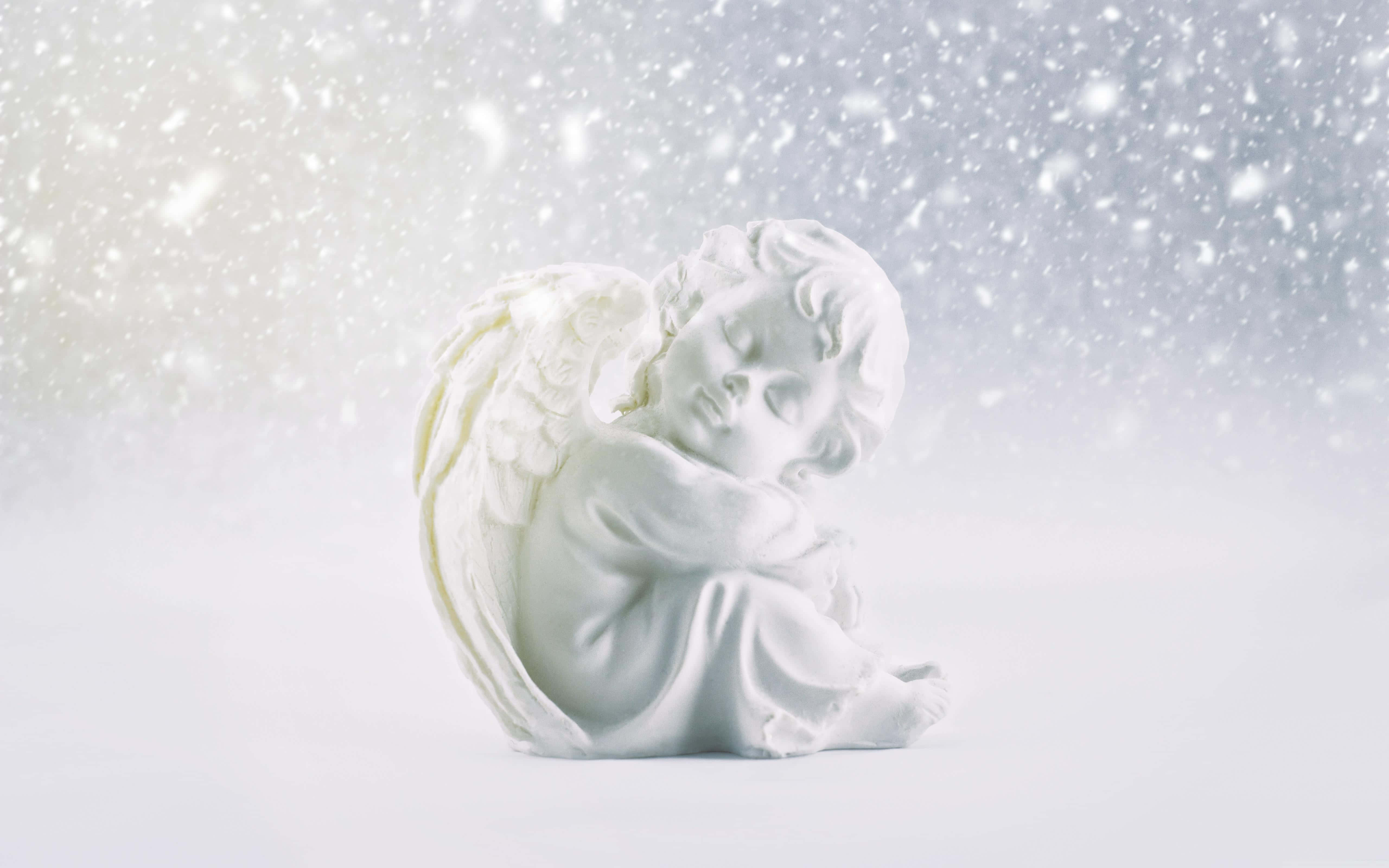 Snow Angel 桌布 壁紙 — 一座寧靜的白色天使雕像蜷縮著安詳入睡，溫柔的雪花在柔和發光的冬日天空中飄舞。