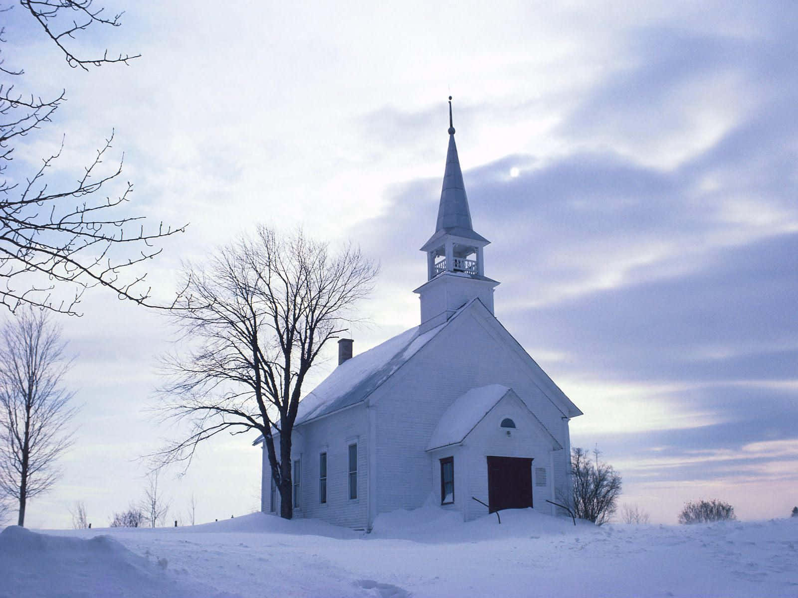 Winter Scene 桌布 壁紙 — 一座寧靜的白色教堂帶有尖頂，獨自矗立在雪覆蓋的田野中，在柔和的暮色天空下。