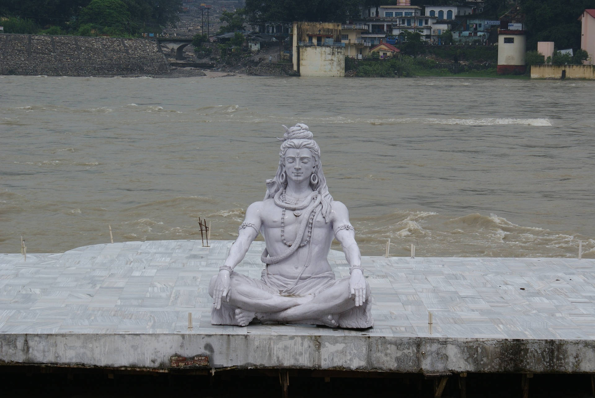 shiv shankar 桌布 壁紙 — 一座寧靜的白色石像，一位冥想的神祇盤腿坐在瓷磚平台上，俯瞰著湍急的棕色河流，背景是一個村莊。