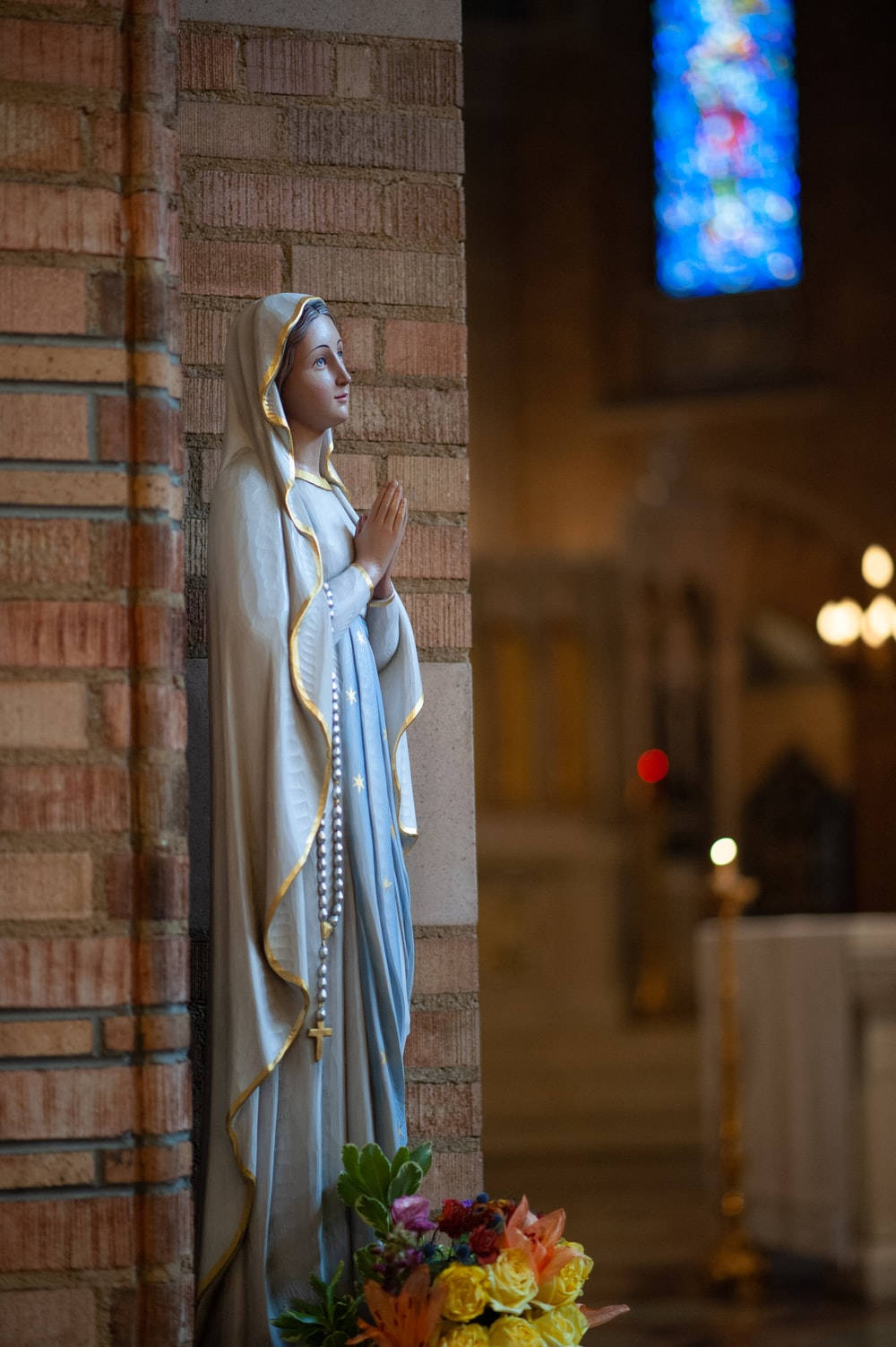 Catholic 桌布 壁紙 — 一座寧靜的聖母瑪利亞祈禱雕像，立於磚柱旁，教堂內光線柔和，底部有彩繪玻璃和鮮花。