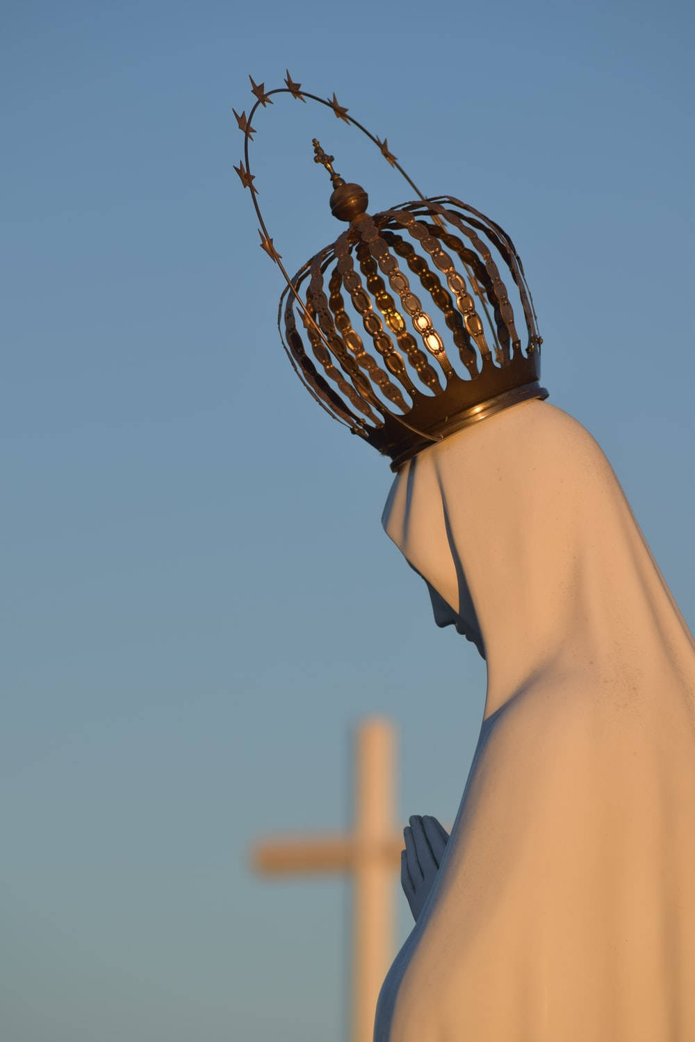 Catholic 桌布 壁紙 — 一座寧靜的聖母瑪利亞雕像，頭戴金色皇冠和星形光環，沐浴在溫暖的夕陽下，背景是清澈的藍天，背後有一個柔和焦點的十字架。