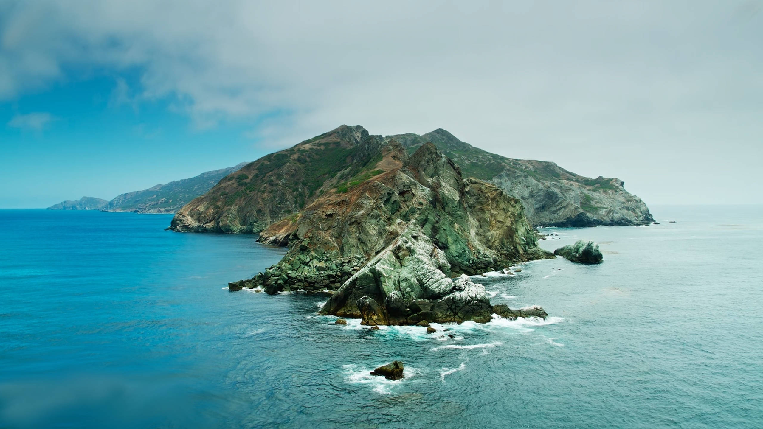Catalina 桌布 壁紙 — 一座崎嶇的岩石島嶼從碧綠色的海水中升起，天空部分多雲，溫和的海浪拍打著它的底部。