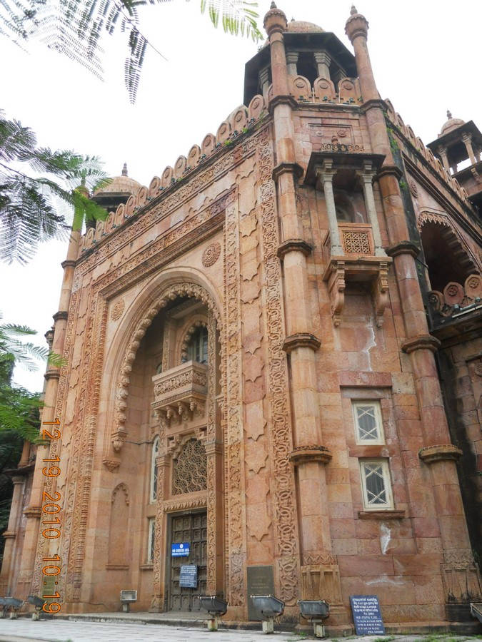 Chennai 桌布 壁紙 — 一座巨大、雕刻精細的粉紅色砂岩建築，帶有圓頂和拱門，矗立在多雲的天空下，左上角有棕櫚葉框住。