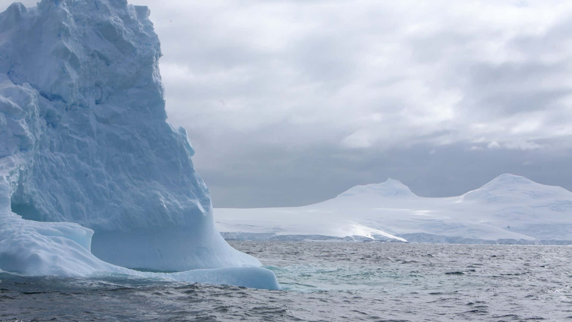 Antarctica 桌布 壁紙 — 一座巨大的冰山，帶有藍色色調的冰塊，聳立在波濤洶湧的灰色水域上，天空多雲，遠處有積雪覆蓋的山脈。