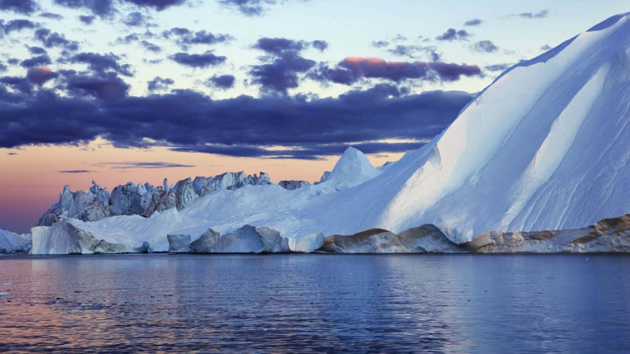 Greenland 桌布 壁紙 — 一座巨大的冰山，擁有鋸齒狀的山峰和光滑的斜坡，在帶有紫色雲彩和橙色地平線的暮色天空下閃耀。