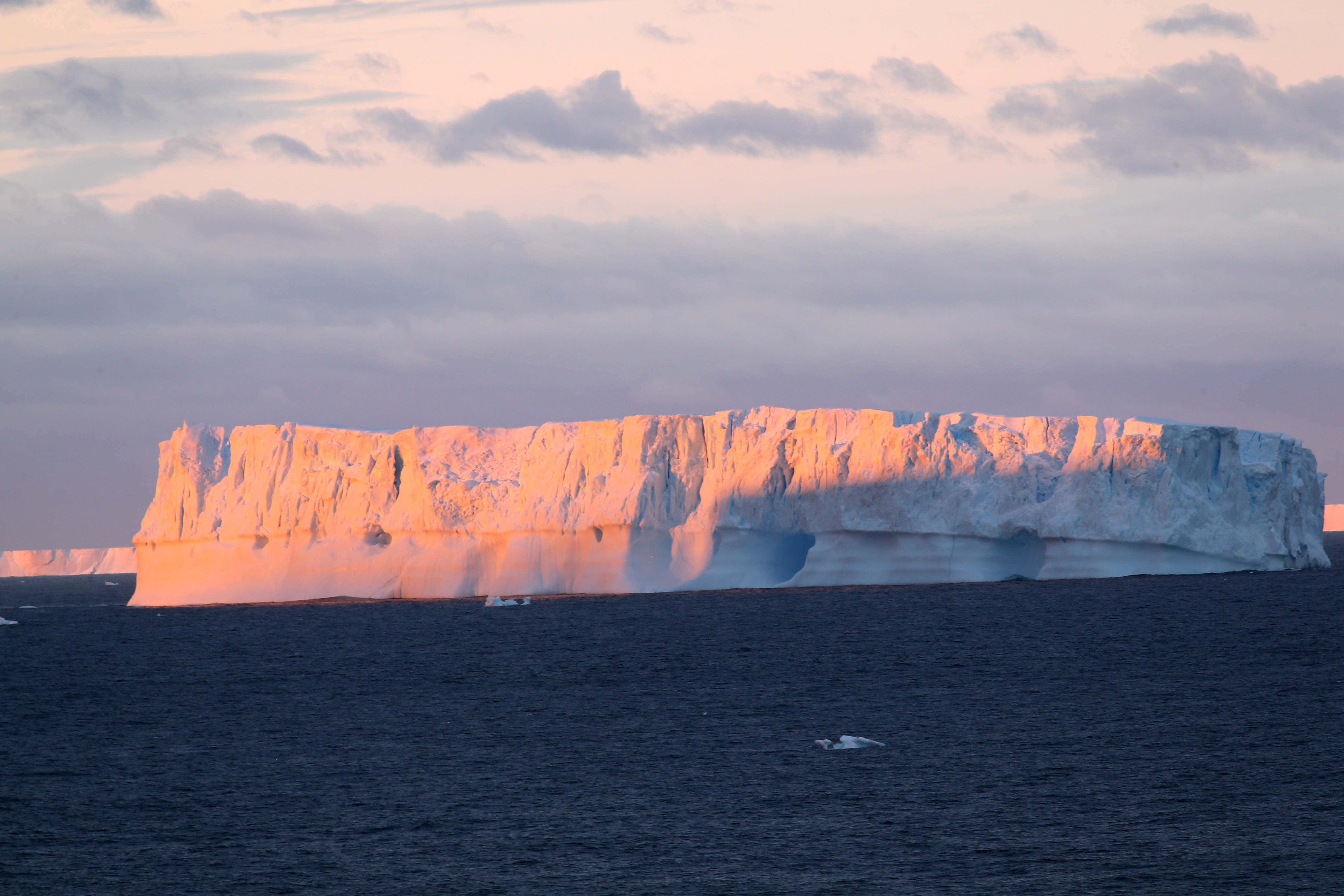 Antarctica 桌布 壁紙 — 一座巨大的冰山在日落時分，在陰暗海洋上方的多雲天空中散發出溫暖的粉紅色光芒。
