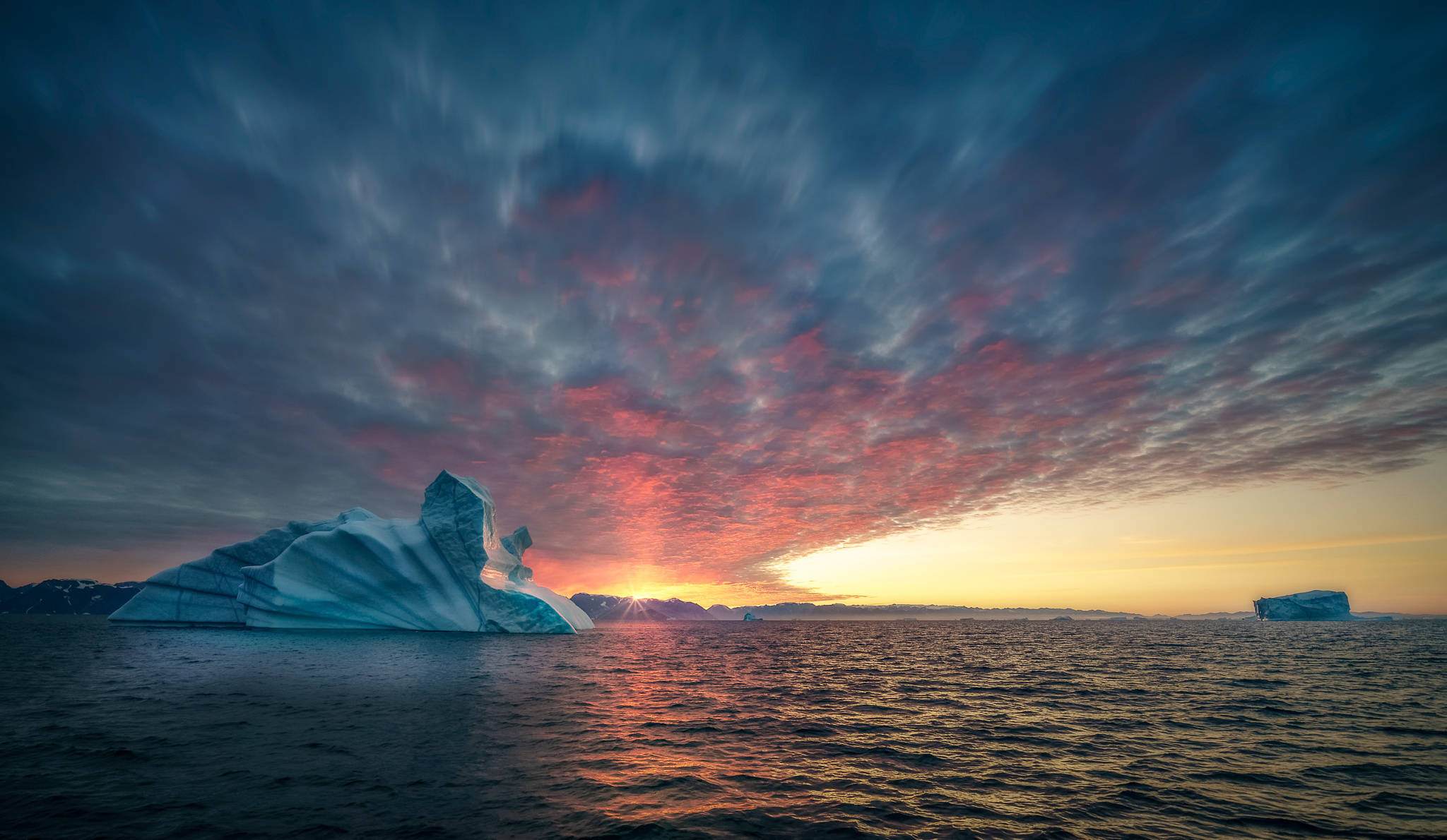 Greenland 桌布 壁紙 — 一座巨大的冰山在火紅的日落天空下發光，天空佈滿粉紅色和橙色的雲彩，映照在平靜的海面上。