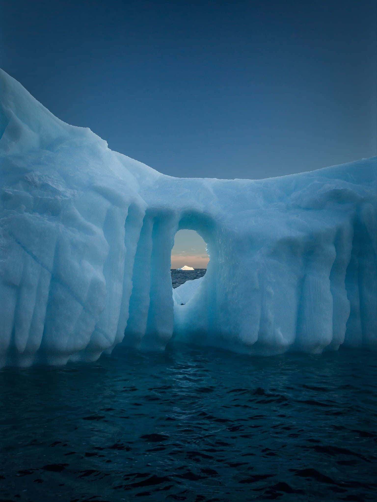 Greenland 桌布 壁紙 — 一座巨大的冰山帶有天然拱門，在深藍色天空下，框住了黑暗海洋上的遠方日落。