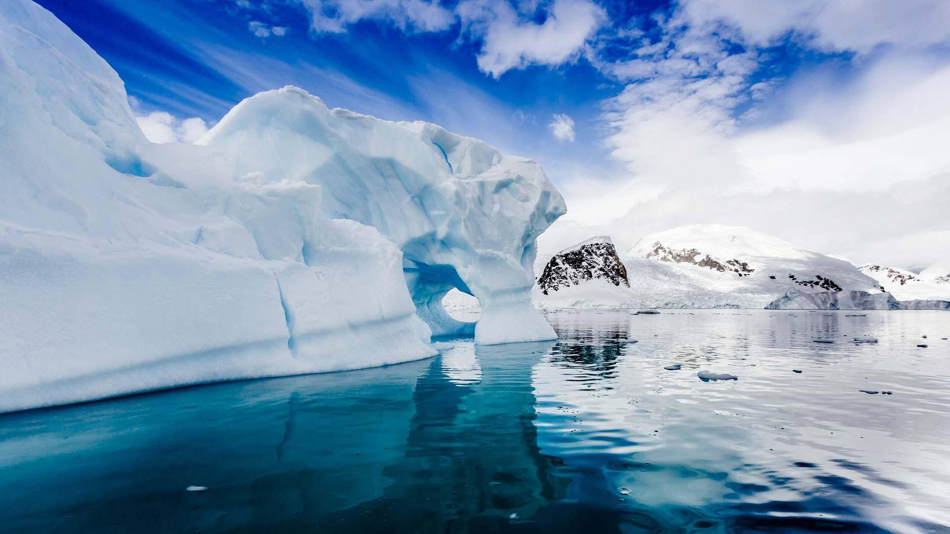 Antarctica 桌布 壁紙 — 一座巨大的冰山帶有天然拱門，矗立在平靜的藍綠色海水中，上方是湛藍的天空，點綴著幾朵白雲。