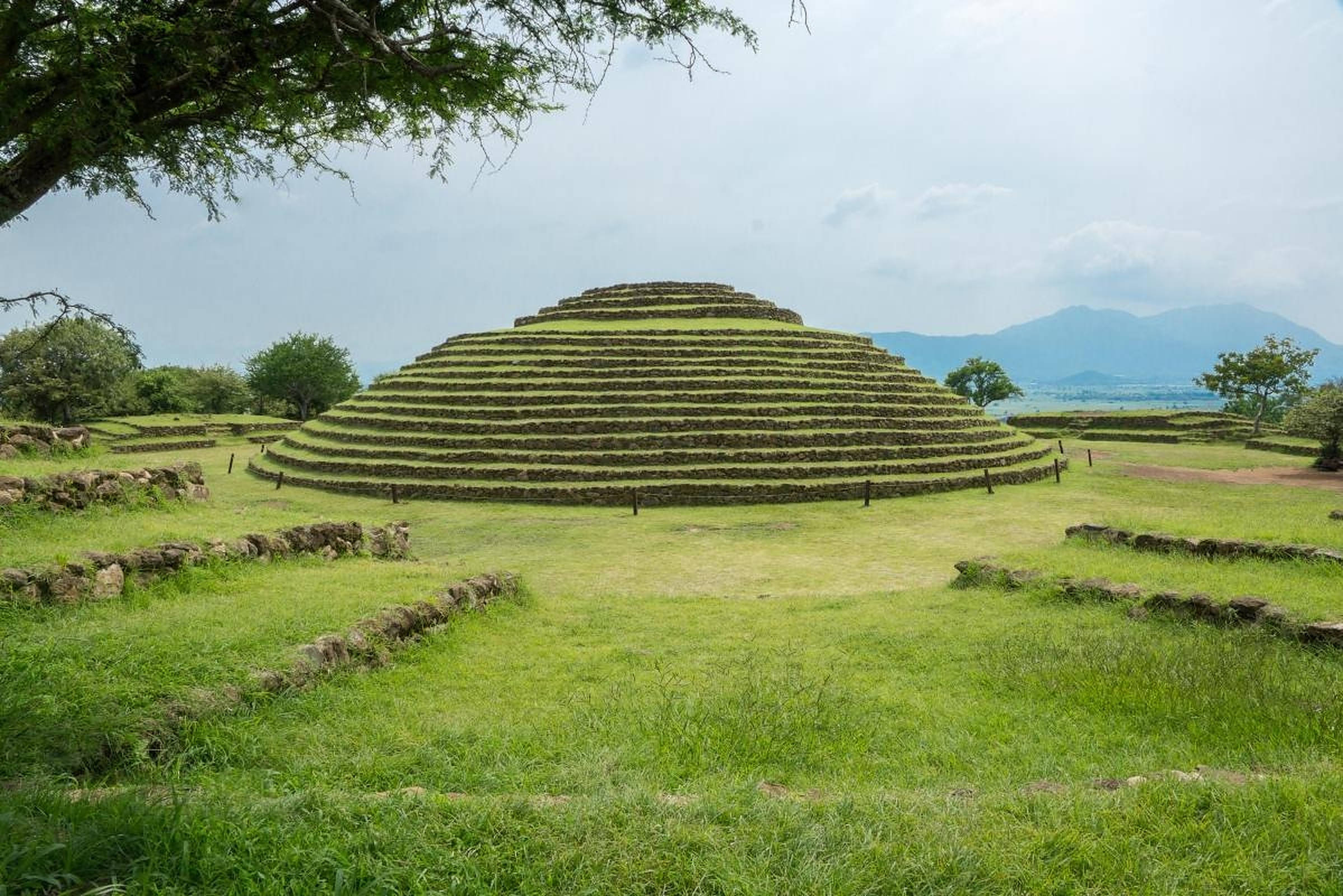 Guadalajara 桌布 壁紙 — 一座巨大的古老階梯金字塔，覆蓋著充滿活力的綠草，矗立在寧靜的開闊田野中，天空多雲，遠處有山脈。