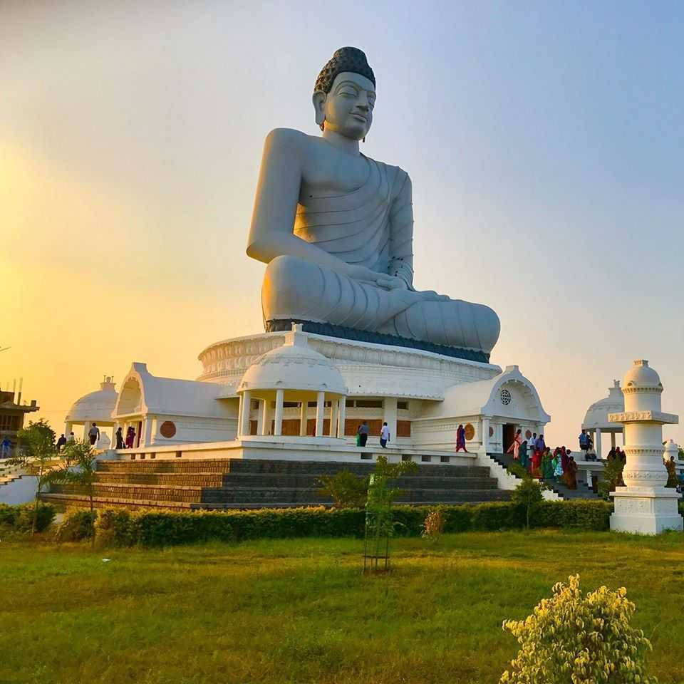 Hyderabad 桌布 壁紙 — 一座巨大的寧靜佛像坐落在白色寺廟建築群頂部，沐浴在金色的日落光芒中，附近有遊客走動。