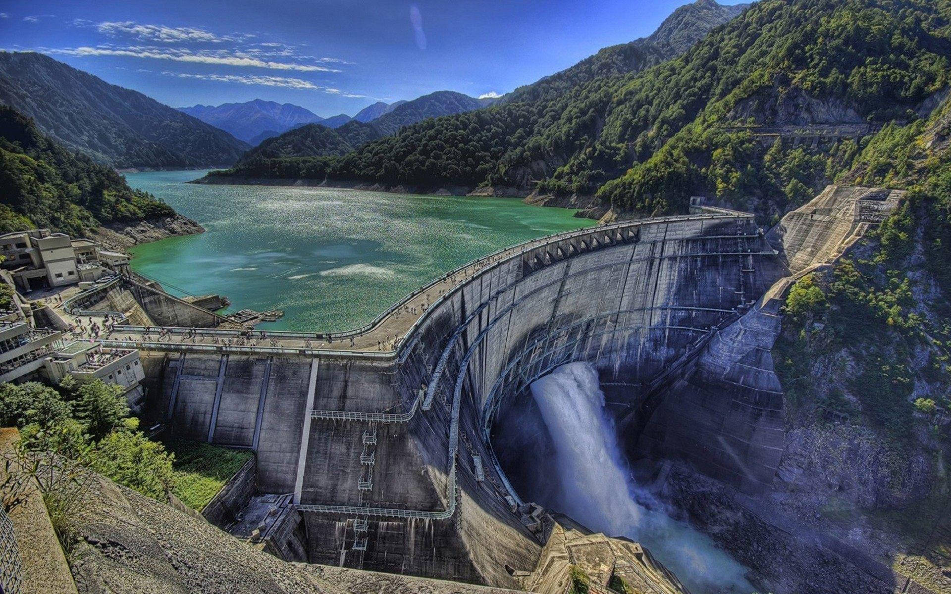 Lake 桌布 壁紙 — 一座巨大的弧形混凝土水壩在明亮的藍天下，將強勁的瀑布洩入翠綠群山環抱的藍綠色水庫中。