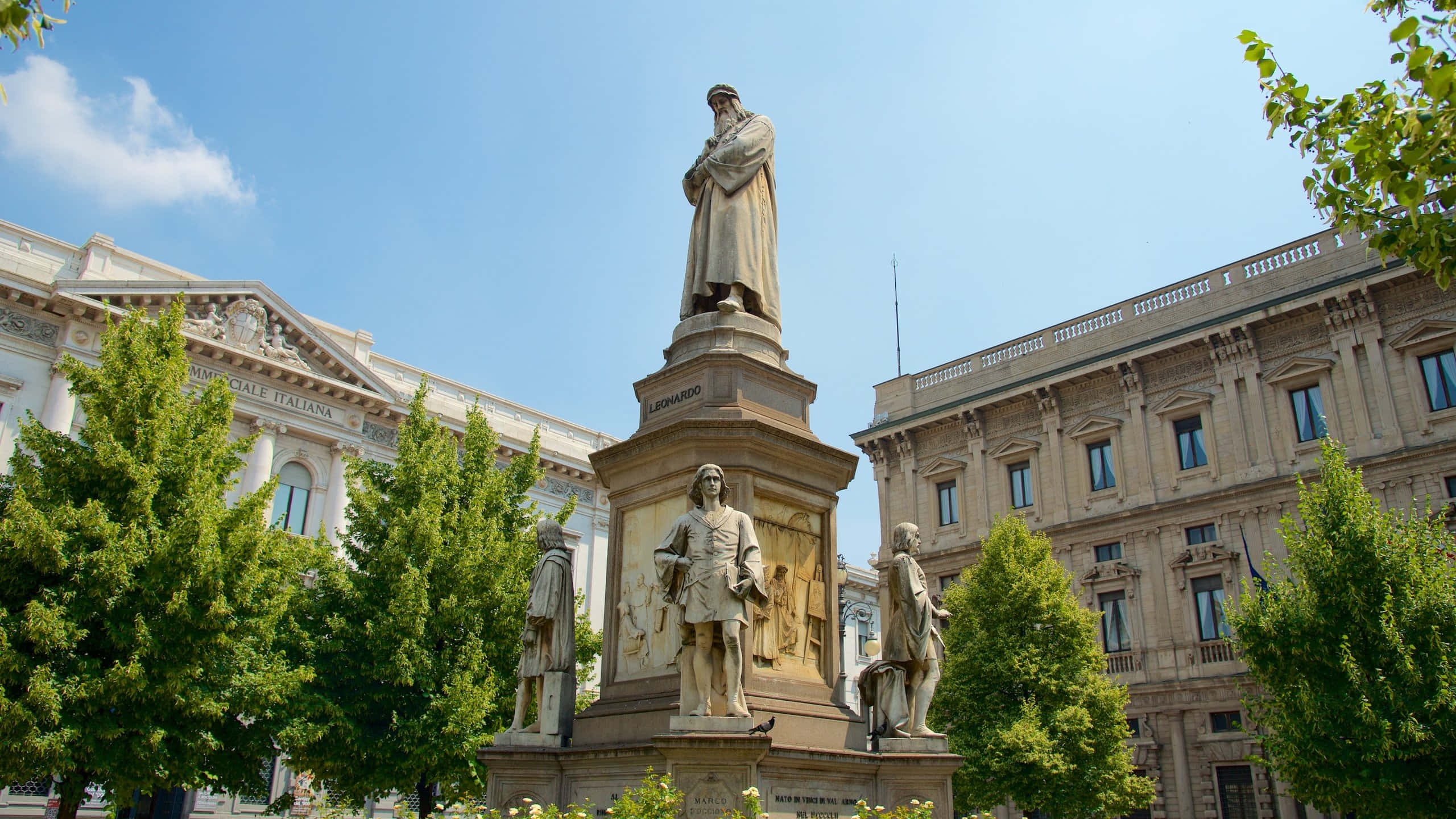 statue 桌布 壁紙 — 一座巨大的李奧納多·達文西石像矗立在陽光明媚的廣場上，周圍環繞著茂密的綠樹和古典建築。